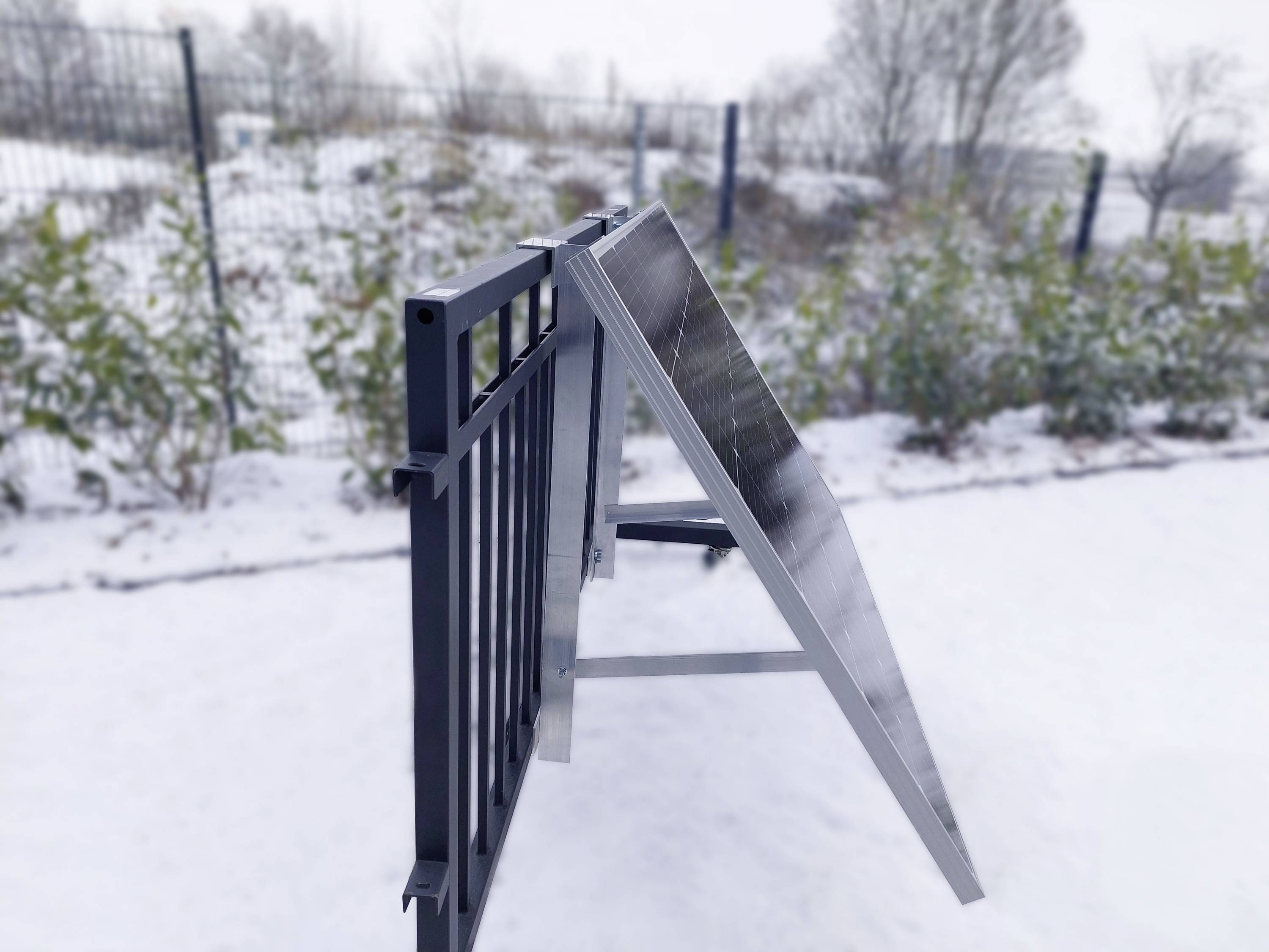 Panel słoneczny jest zamontowany pod skosem na stelażu. W tle widoczny jest płot i pokryte śniegiem rośliny.