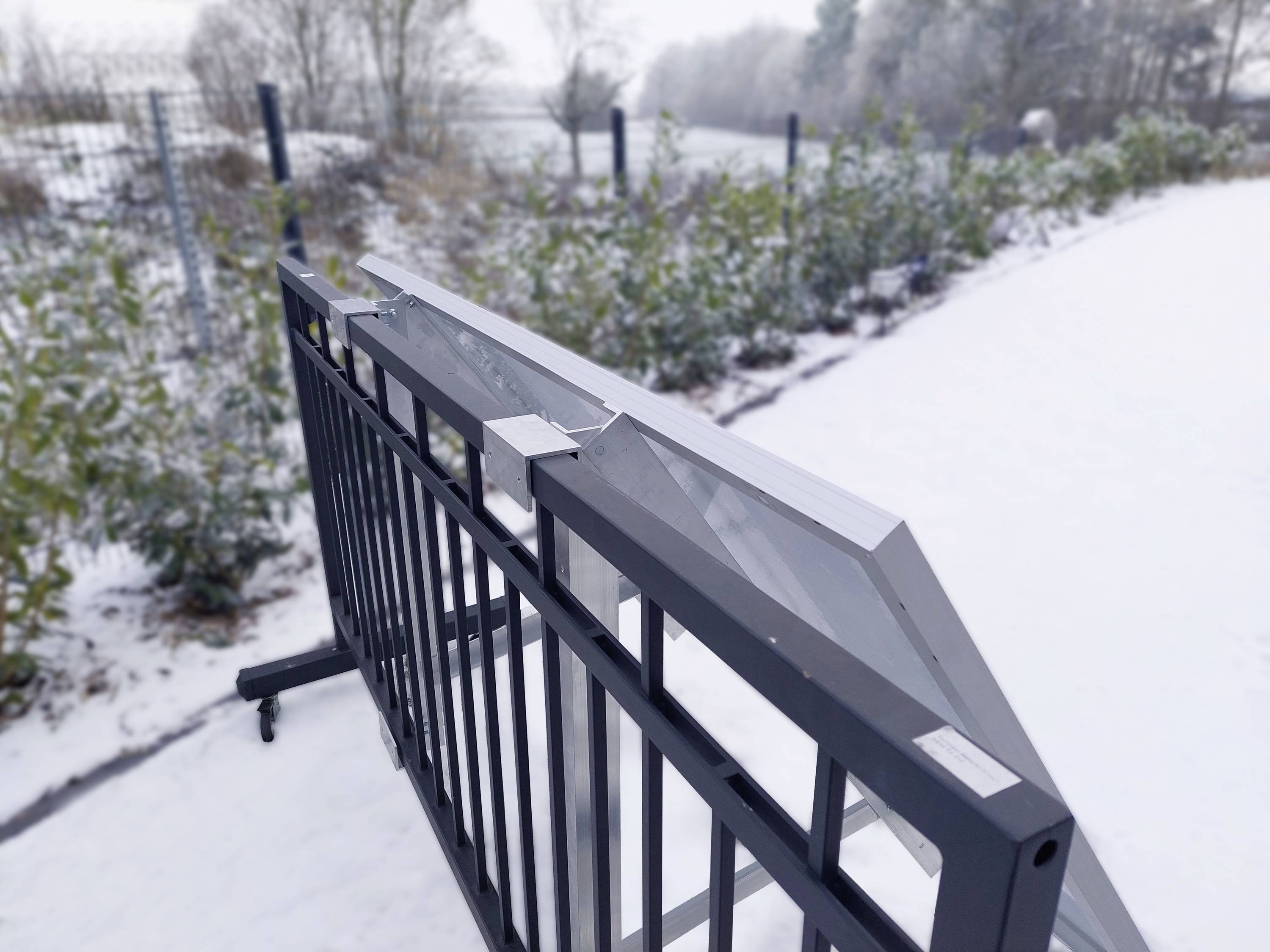 'Składana czarna bariera stoi w zaśnieżonej przestrzeni zewnętrznej, otoczona roślinami i płotem.'