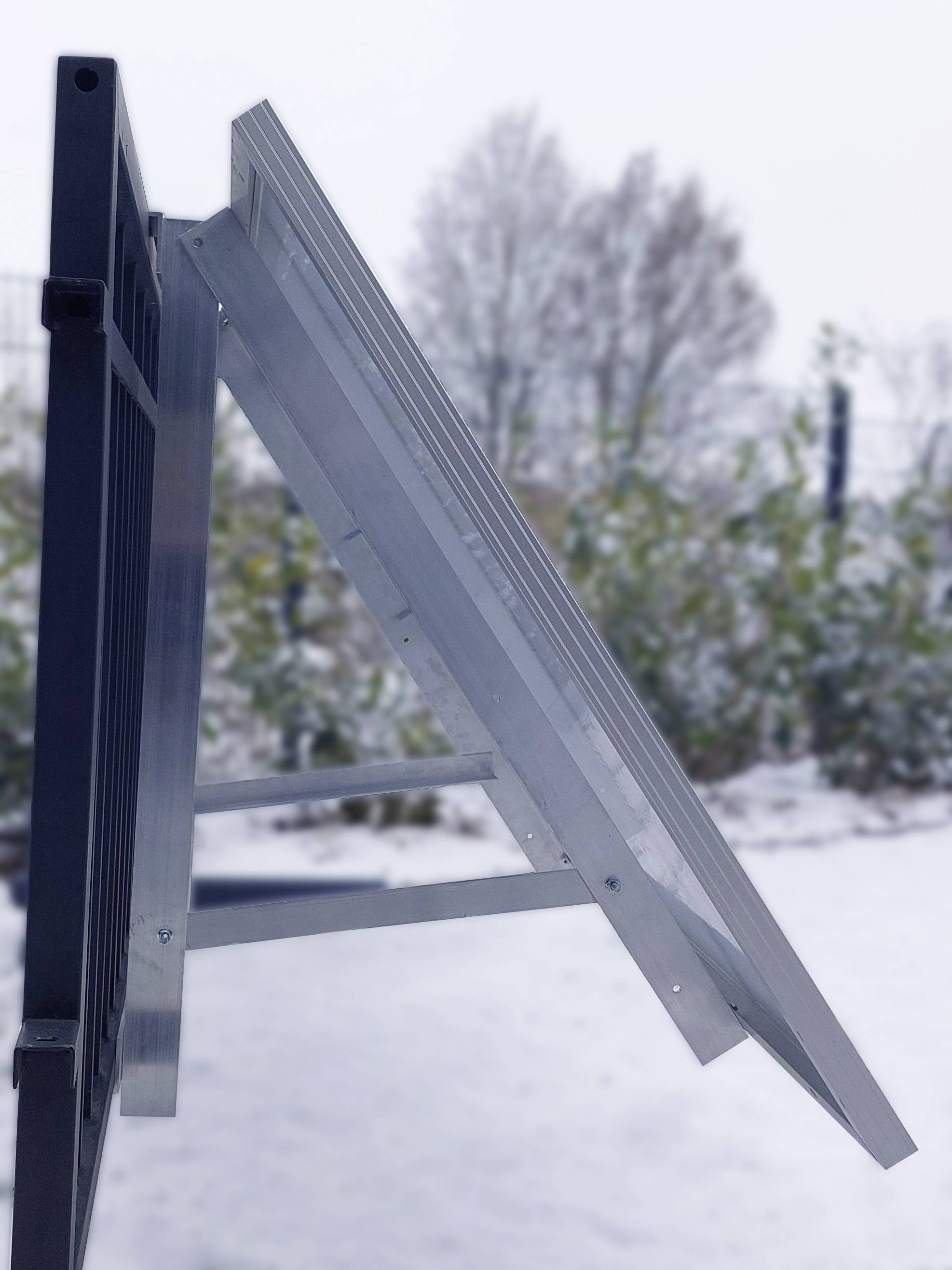 Panel słoneczny jest zamontowany skośnie na metalowej ramie, ustawionej na wolnym powietrzu. W tle widoczna jest zaśnieżona ziemia i drzewa.