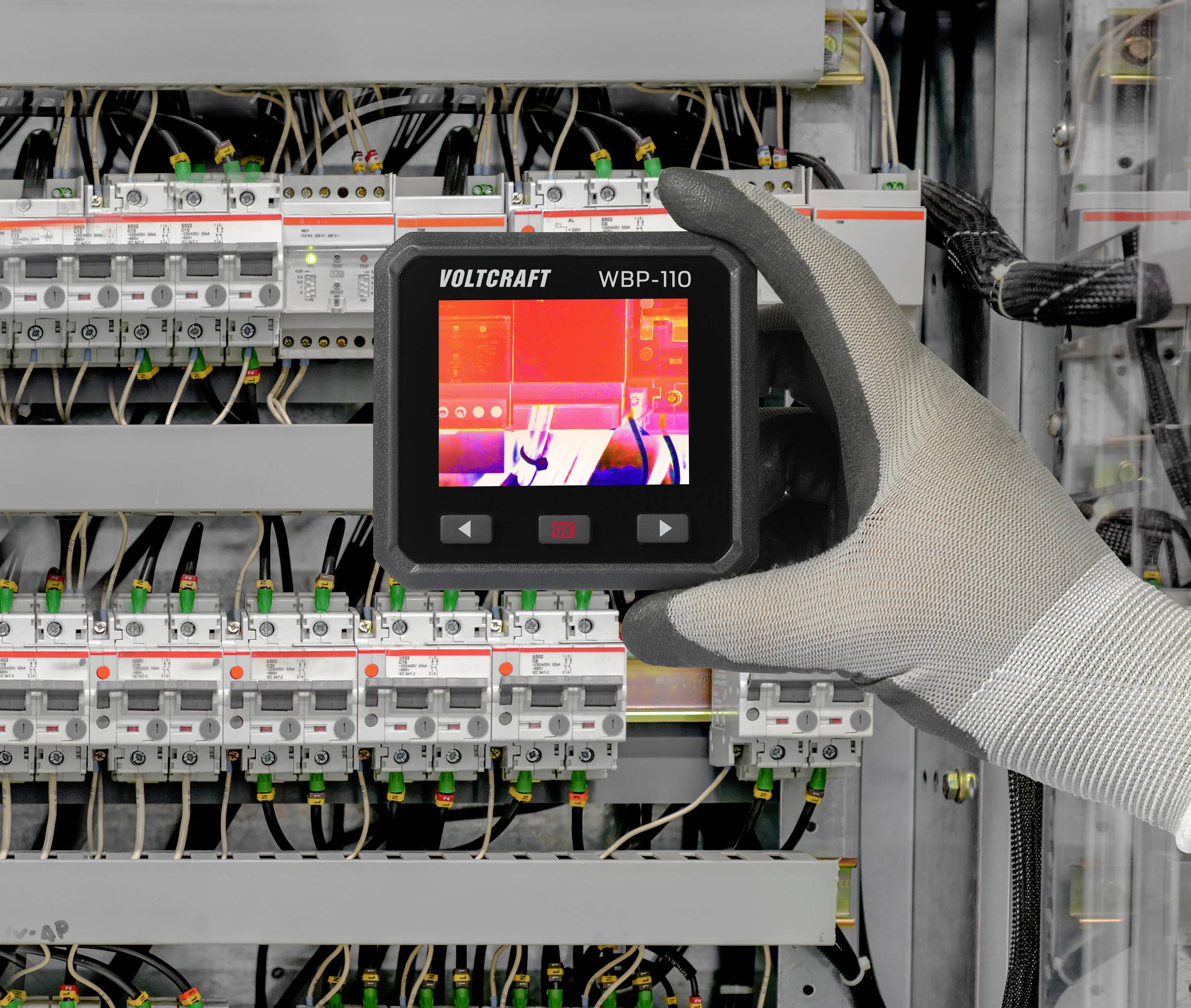 Rękawica trzyma kamerę termowizyjną przed rozdzielnicą elektryczną. Ekran wyświetla termiczny obraz obwodów elektrycznych.