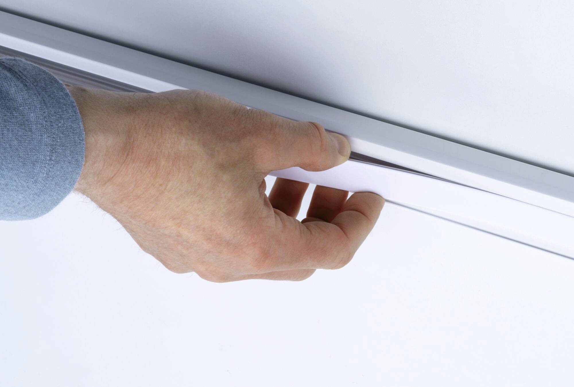 Dłoń w niebieskiej koszuli przesuwa białą płytę lub przesłonę do wąskiej prowadnicy przy ścianie lub suficie.