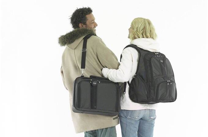 Kobieta i mężczyzna stoją uśmiechnięci plecami do siebie. Kobieta ma na sobie duży czarny plecak, a mężczyzna czarną torbę na ramieniu.