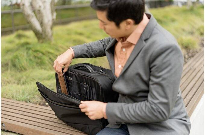 Mężczyzna w szarym garniaku siedzi na ławce i wkłada tablet do czarnej torby na laptopa.