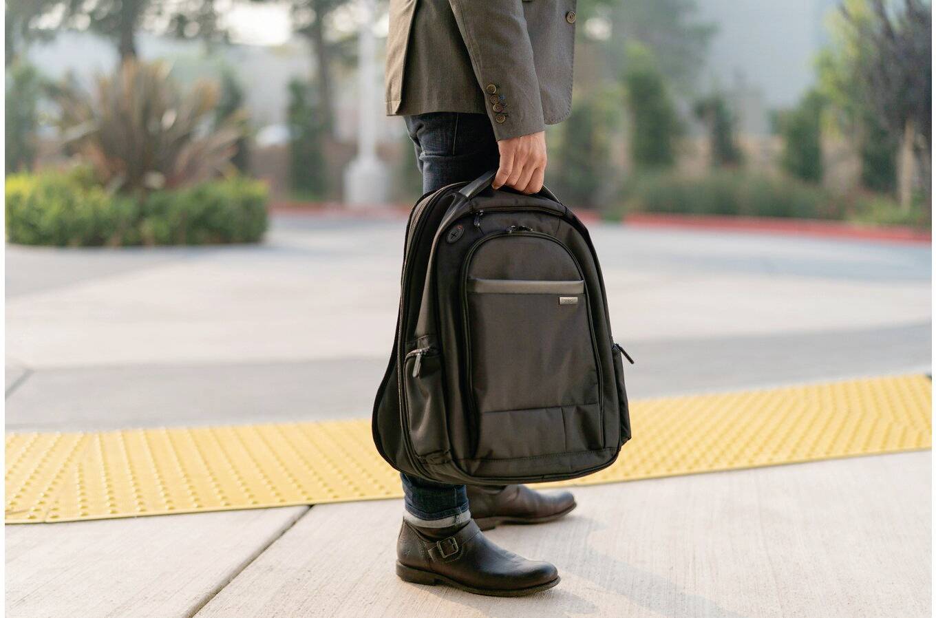 Osoba przechodzi chodnikiem, trzymając czarny plecak w ręku. Tło jest lekko rozmyte.