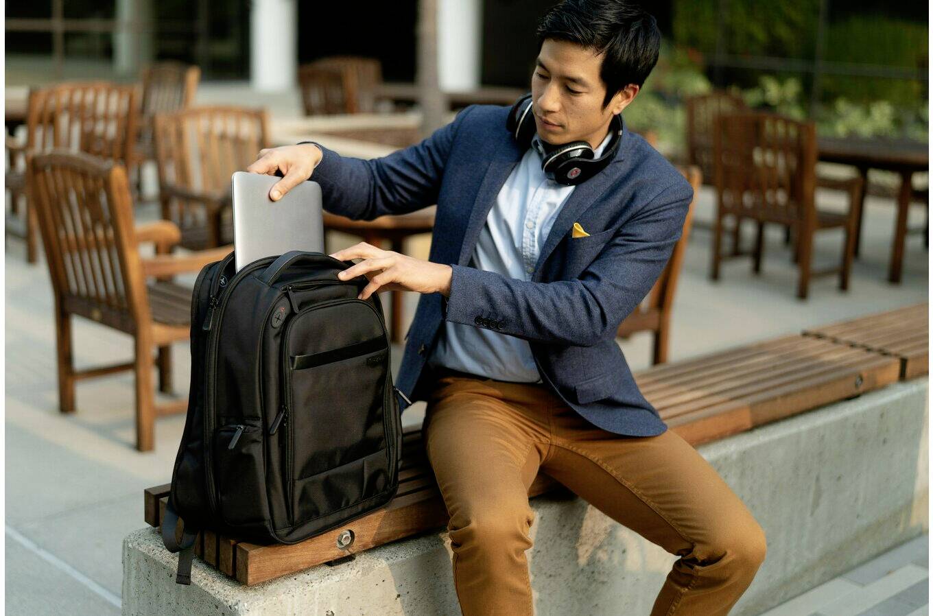 Mężczyzna siada na ławce na wolnym powietrzu i wkłada laptopa do plecaka. Ma słuchawki zawieszone na szyi.