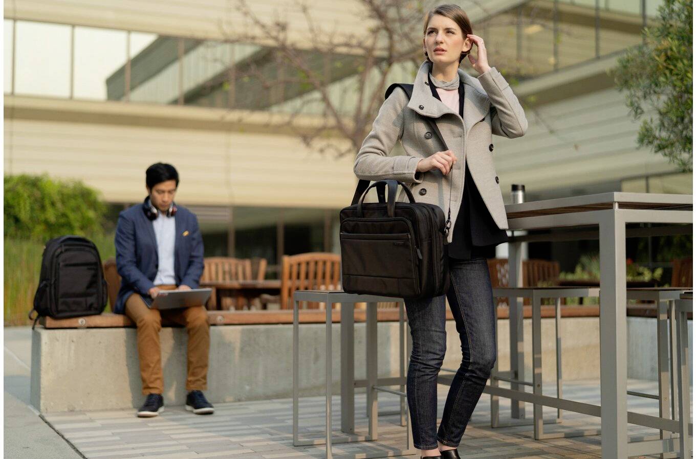 Kobieta stoi na tarasie, trzyma aktówkę i rozmawia przez telefon. W tle mężczyzna siedzi na ławce z laptopem.