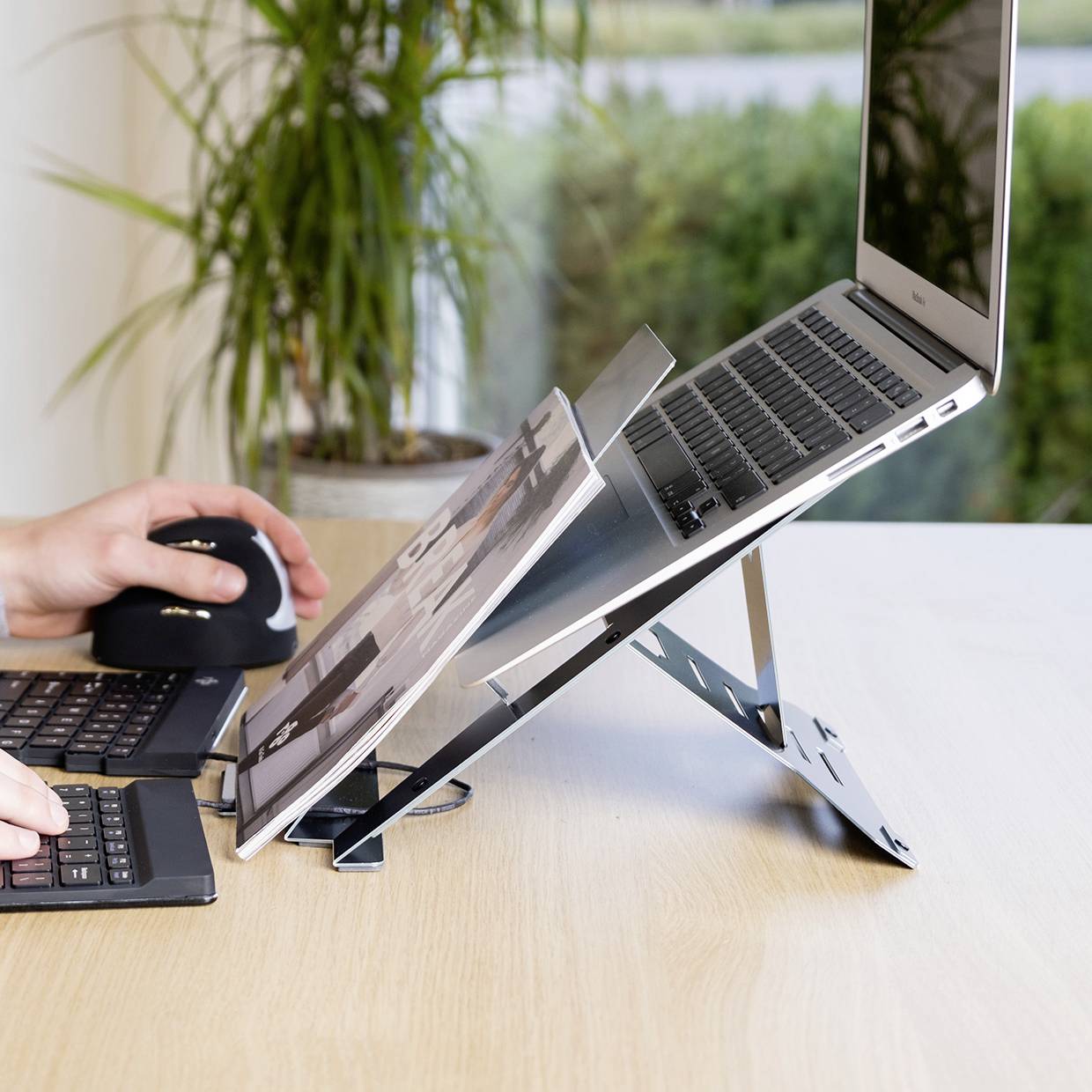 Laptop na stojaku, obok siedzi osoba korzystająca z myszy i klawiatury. W tle widać okno z widokiem na zieleń.