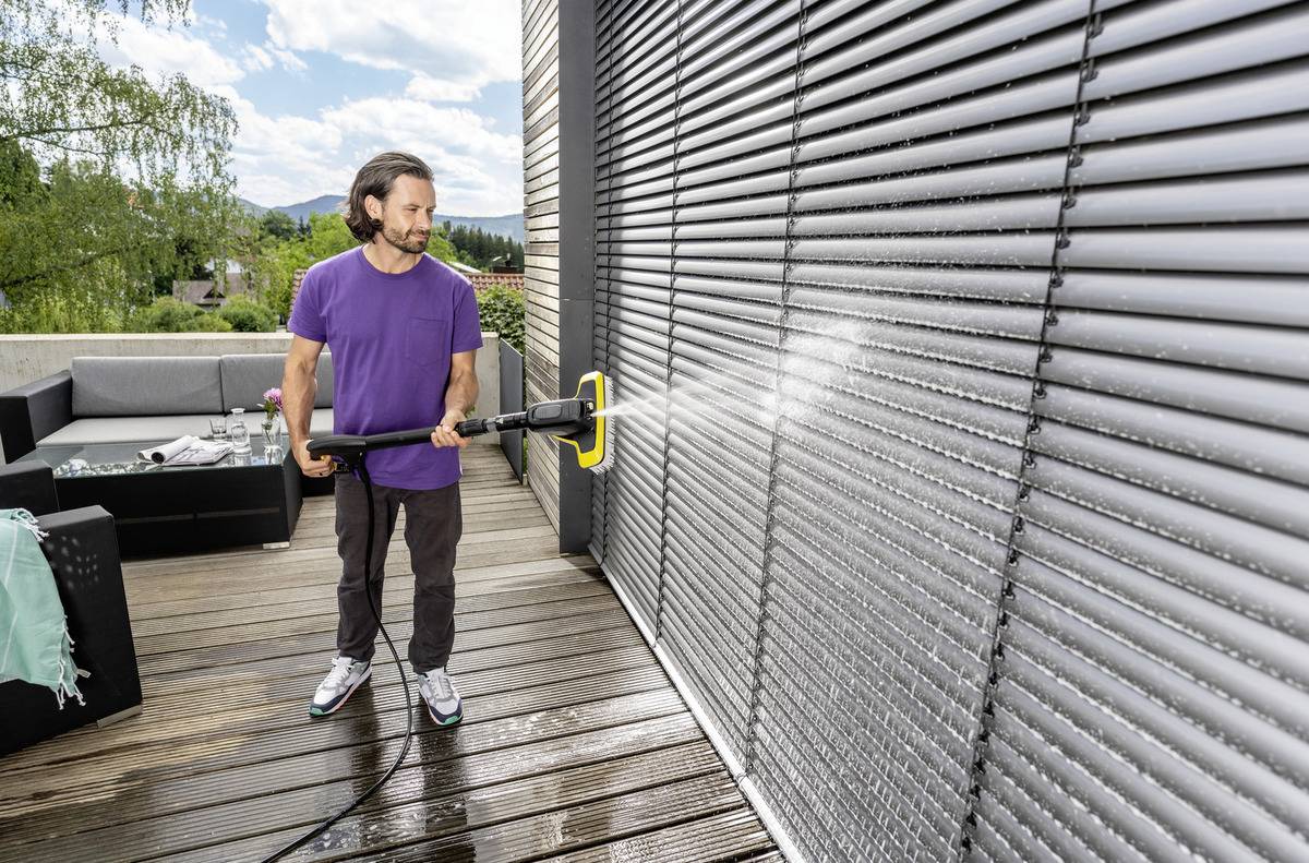 Mężczyzna czyszczący zamknięte żaluzje myjką ciśnieniową na drewnianym tarasie, ubrany w fioletową koszulę i dżinsy. W tle widoczne są meble ogrodowe.