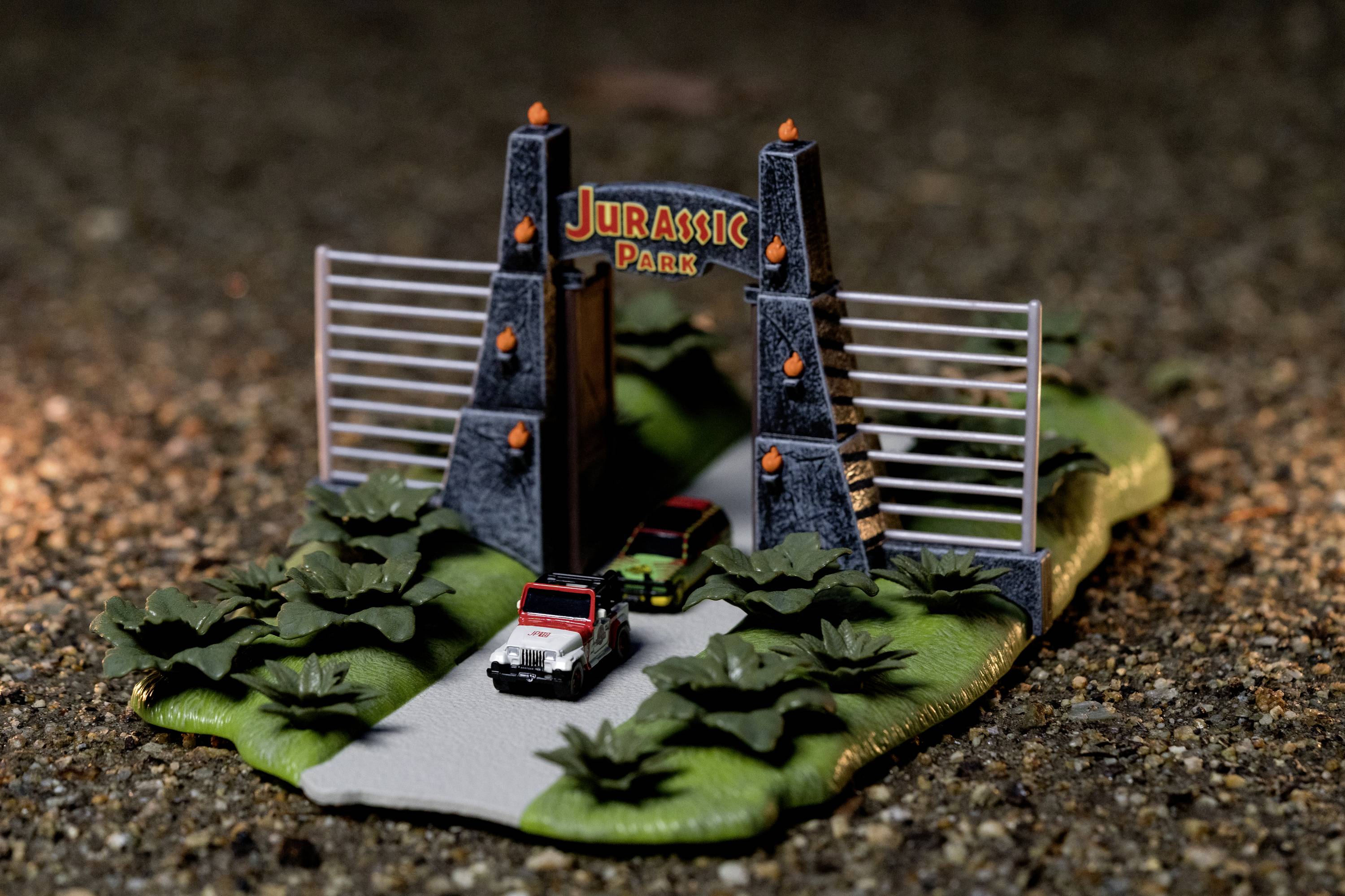 Miniaturowy model Parku Jurajskiego z dwoma pojazdami, bramą i otoczeniem. Pojazdy są zaparkowane w kierunku bramy.