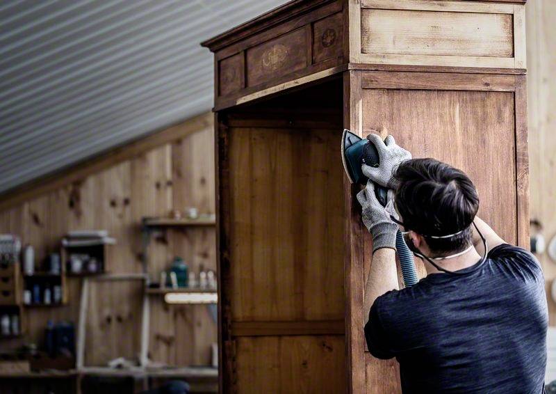 Mężczyzna szlifuje drewnianą szafę w warsztacie. Ma na sobie rękawice robocze i maskę ochronną. W tle widoczne są półki z narzędziami.