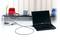 Laptop jest zabezpieczony linką antykradzieżową do biurka. W tle znajdują się krzesła i kolejne laptopy.