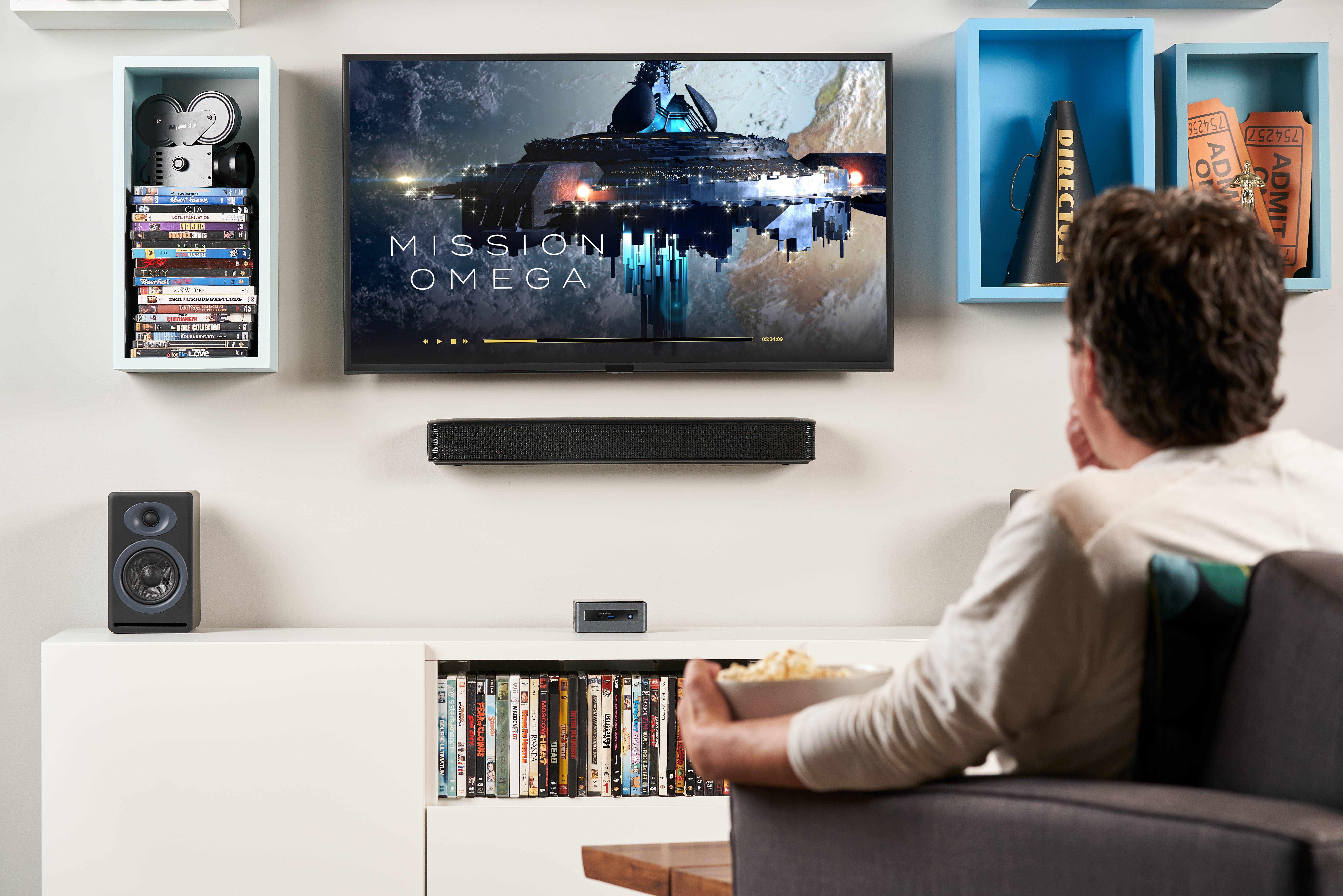 Mężczyzna siedzi na kanapie i ogląda film o tytule 'Misja Omega' w telewizji. W ręku trzyma miskę popcornu.