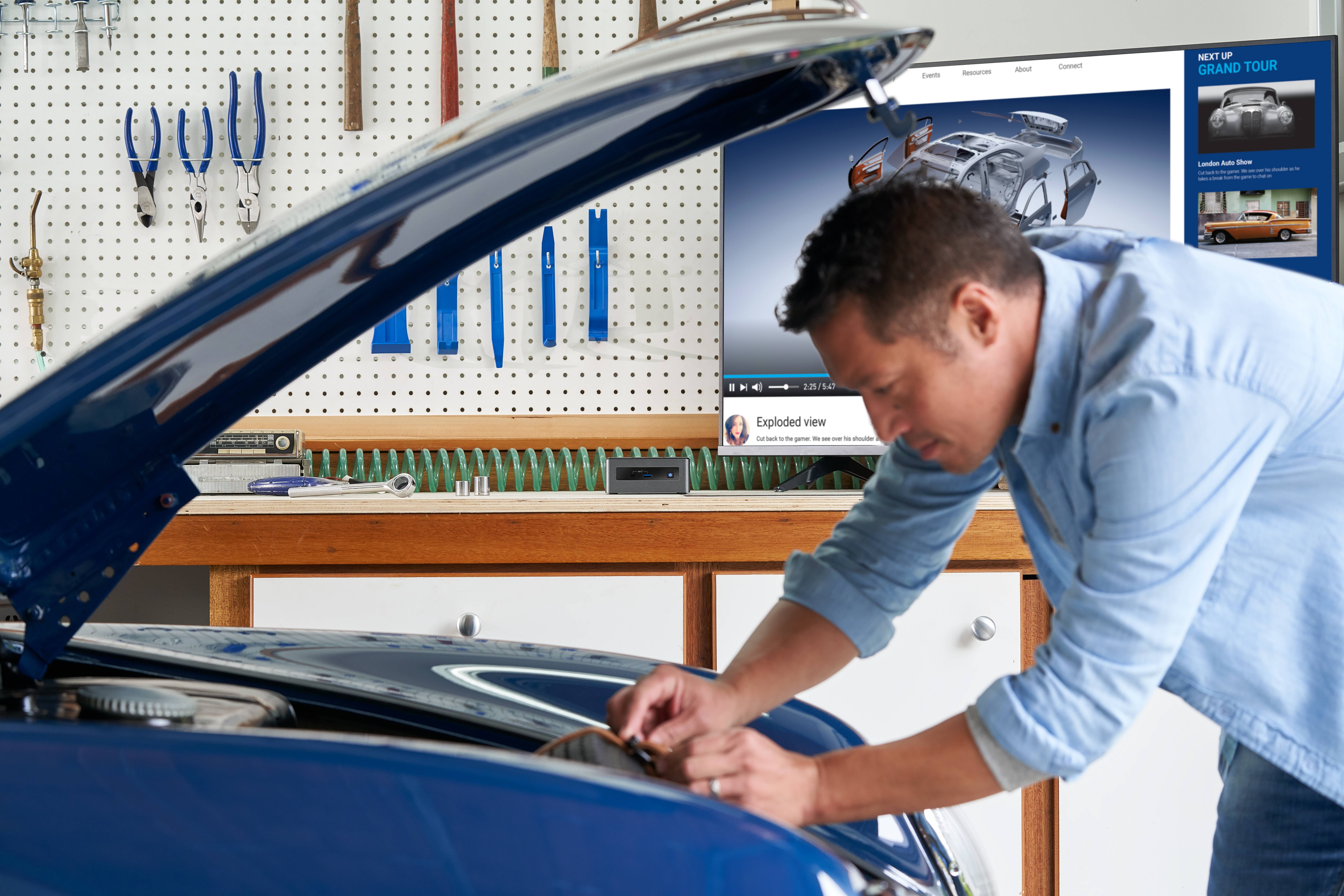 Mężczyzna pracuje przy otwartej komorze silnika samochodu w warsztacie. W tle widoczne są narzędzia oraz ekran z prezentacjami technicznymi.