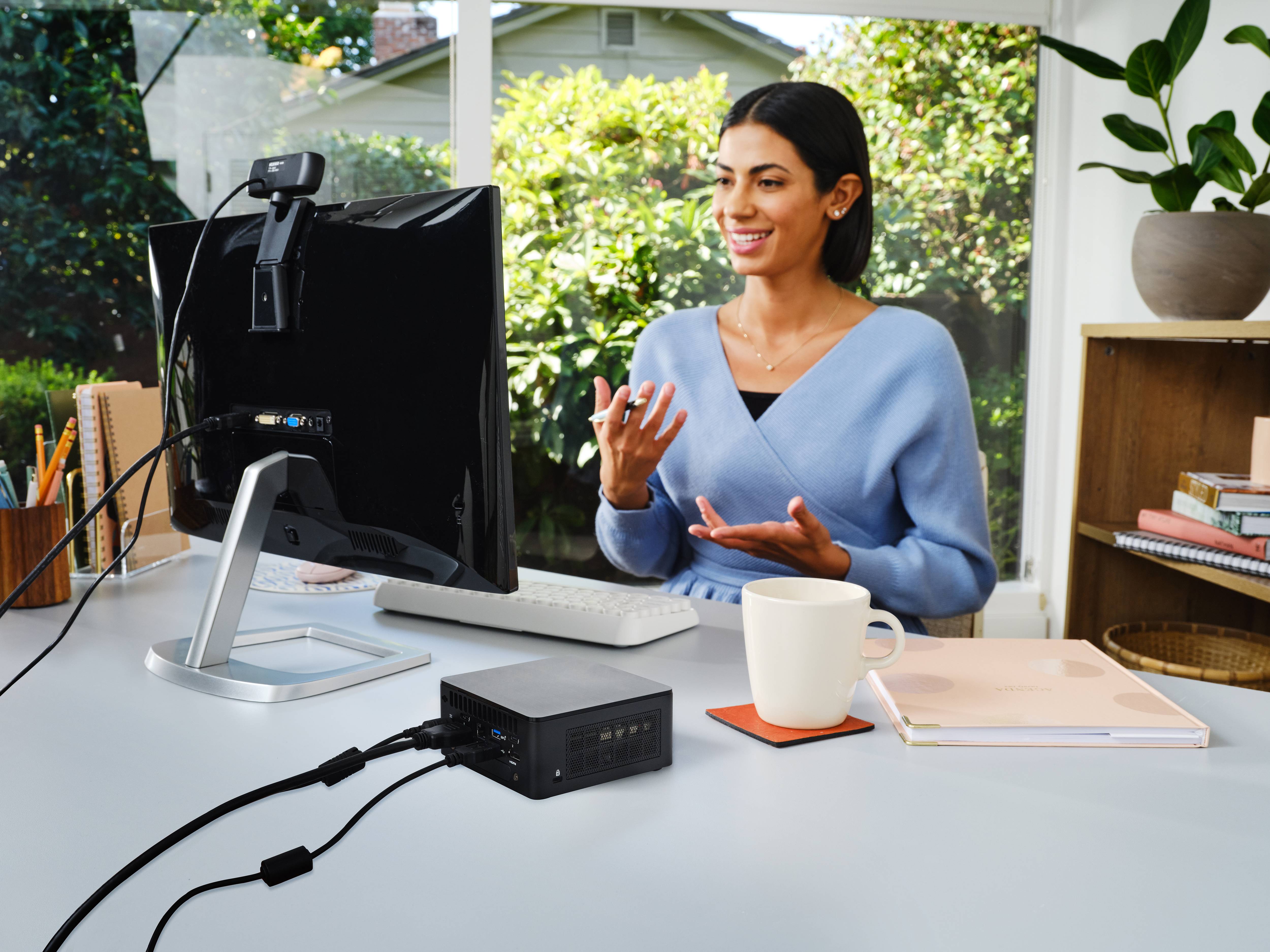 Kobieta uśmiecha się, mówiąc do kamery przed ekranem komputera. Biurko jest wyposażone w książki i akcesoria biurowe.