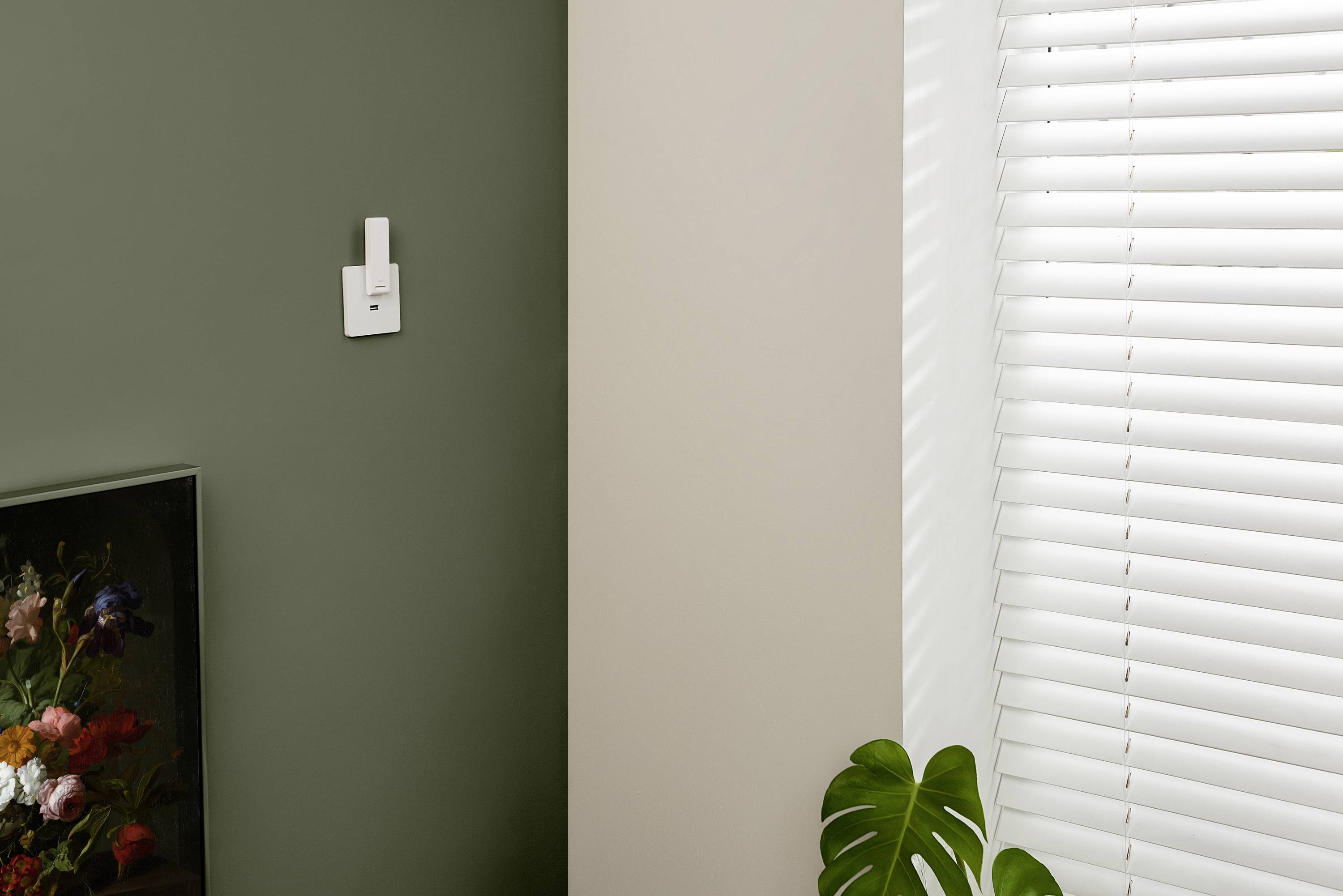 Obraz kwiatowy na oliwkowozielonej ścianie, obok białej żaluzji i rośliny na pierwszym planie.