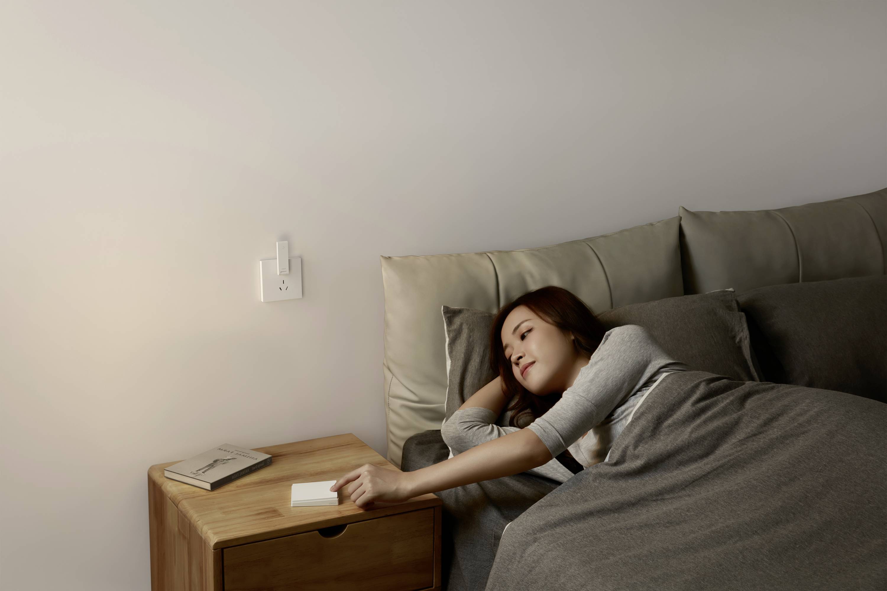 Kobieta leży śpiąca w łóżku, sięgając po budzik stojący na nocnym stoliku. Pomieszczenie wydaje się spokojne i skromnie urządzone.