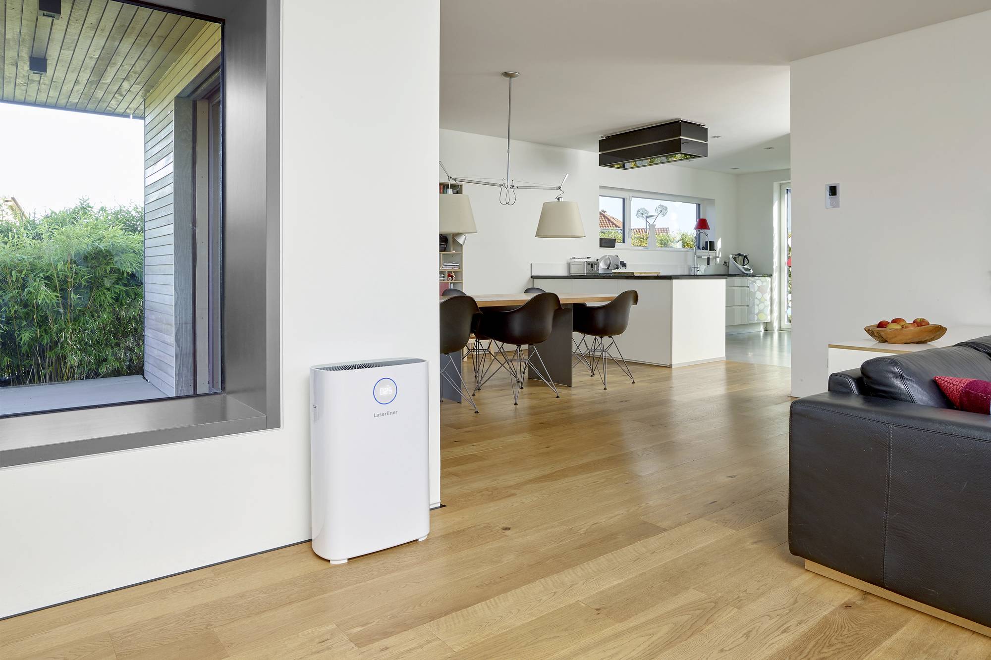 Nowoczesny salon z drewnianą podłogą, szarą sofą, oczyszczaczem powietrza przy białej ścianie i otwartą kuchnią w tle.