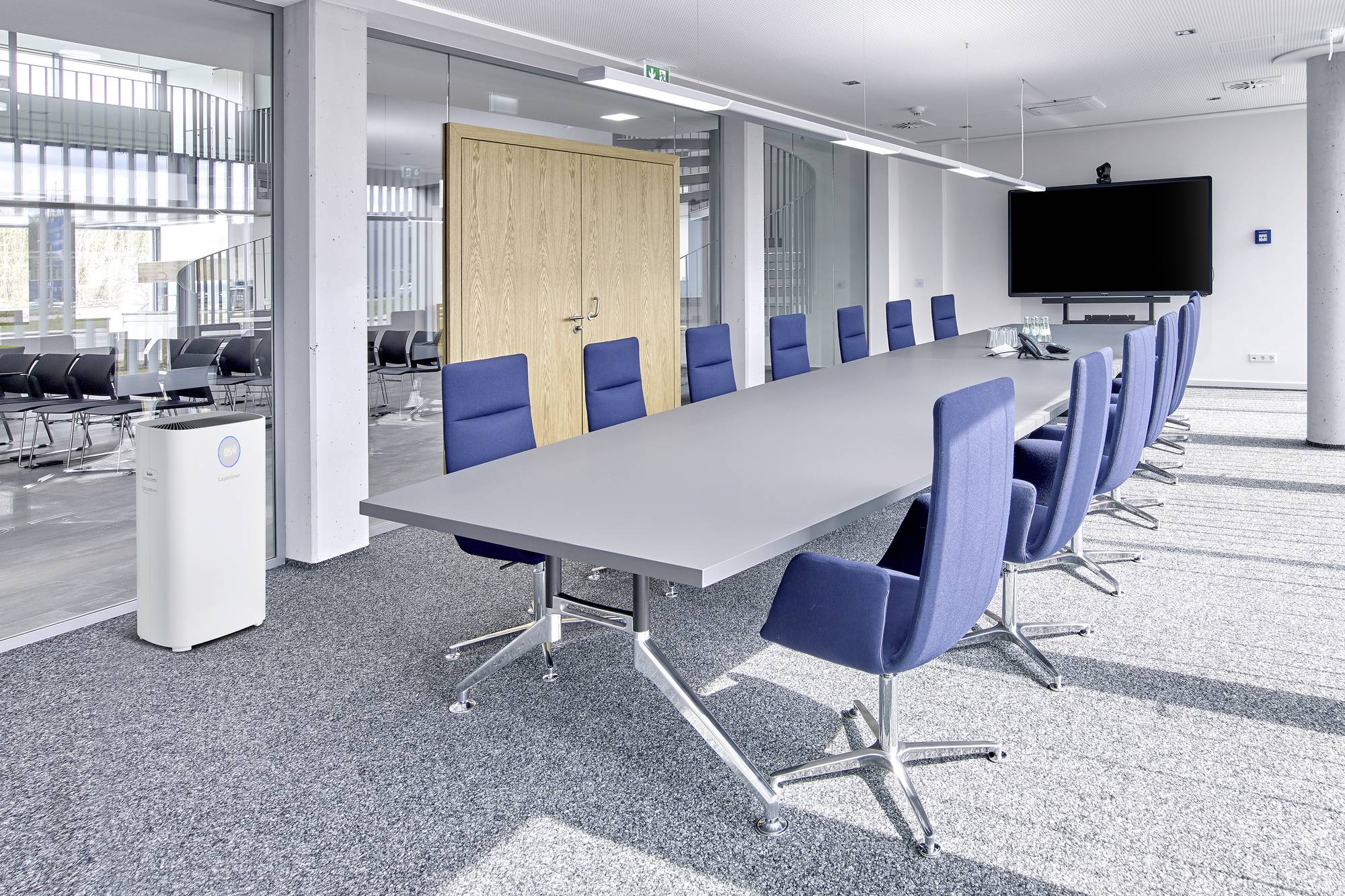 Sala konferencyjna z dużym prostokątnym stołem i niebieskimi krzesłami. Na ścianie wisi duży ekran. Pomieszczenie jest jasne i nowoczesne.