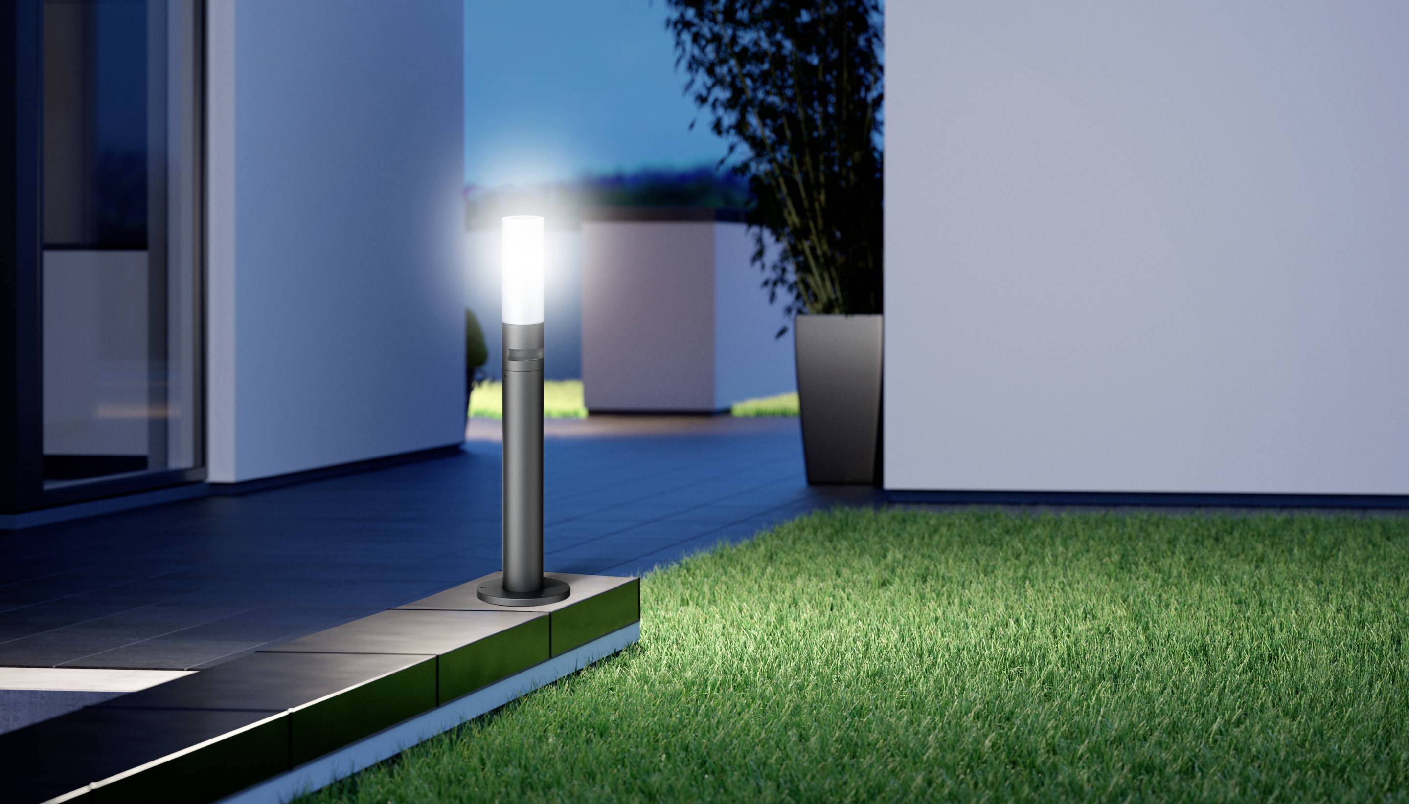 Nowoczesna lampa zewnętrzna oświetla wybrukowaną ścieżkę wzdłuż budynku, otoczoną starannie utrzymanym zielonym trawnikiem.