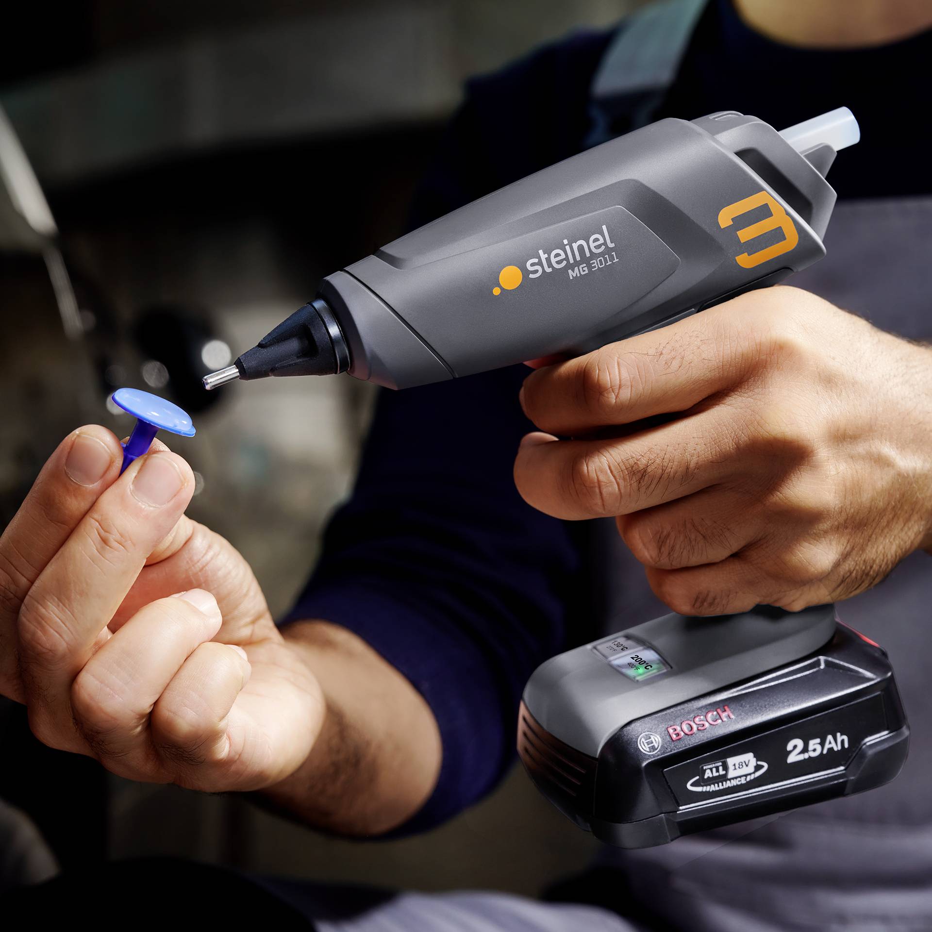 Osoba trzyma pistolet do klejenia na gorąco Steinel MG 3002 i wkłada niebieski wkład kleju do pistoletu. Poniżej leży akumulator Bosch 2,5Ah.