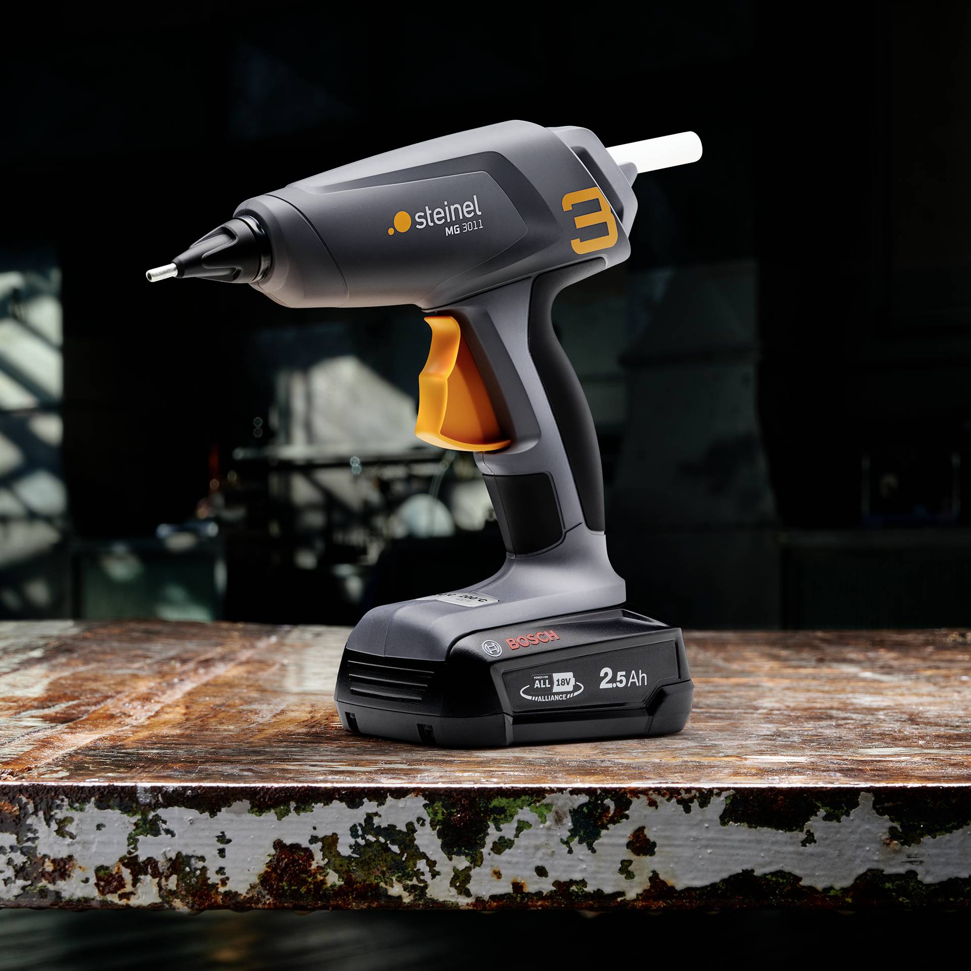 Pistolet do kleju leżący na stole, gotowy do użycia. Pistolet jest czarny z pomarańczowymi i szarymi detalami.