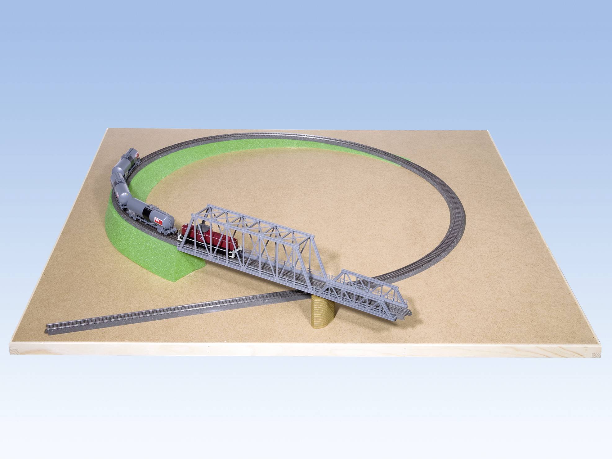 Miniaturowy pociąg jedzie po okrężnej trasie, przejeżdżając przez szary most umieszczony na drewnianej podstawie.