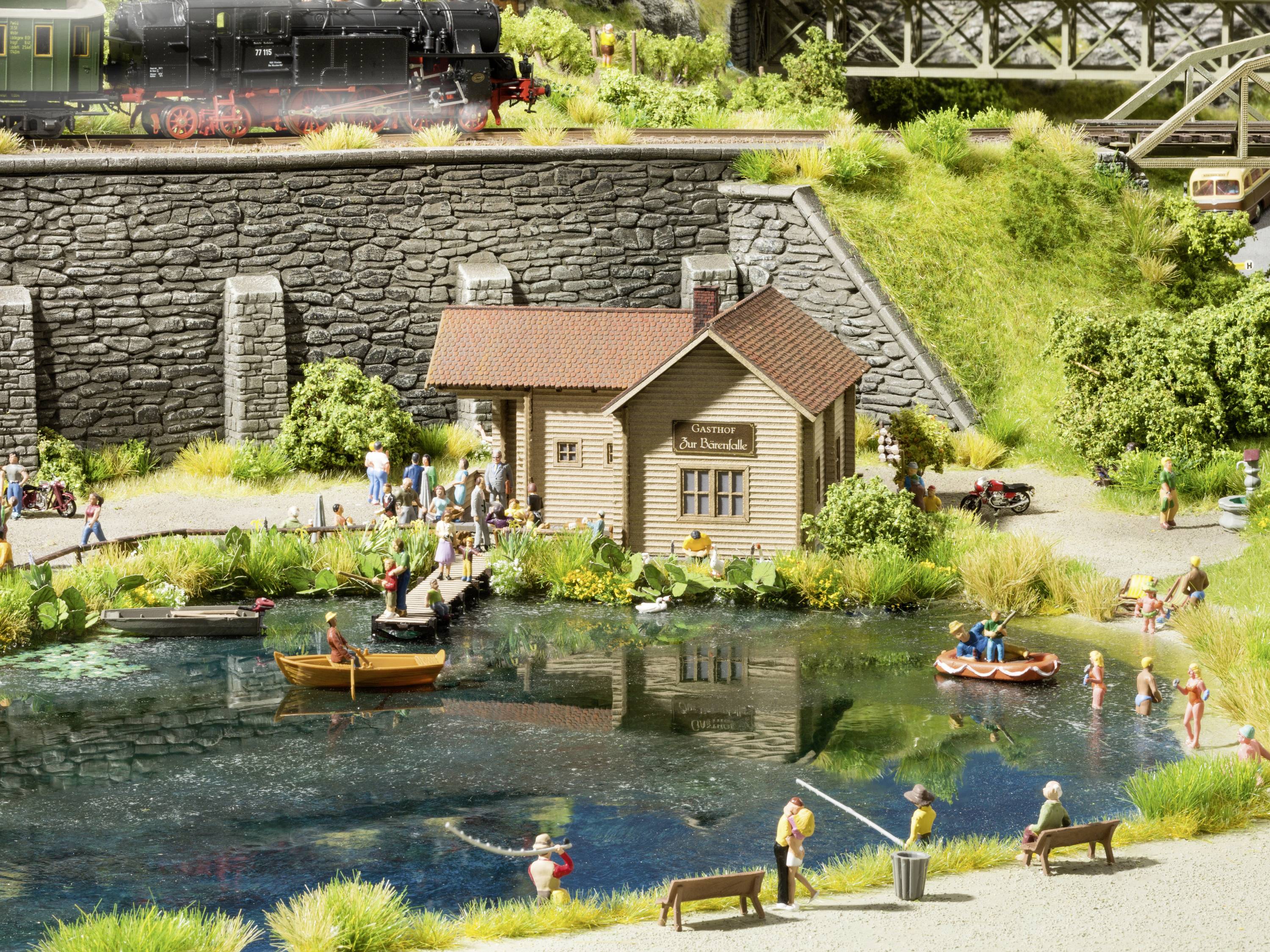 Miniaturowa sceneria z oczkiem wodnym, ludźmi kąpiącymi się, wędkującymi i pływającymi łódką. Budynek z napisem 'Parowóz', most kolejowy w tle.