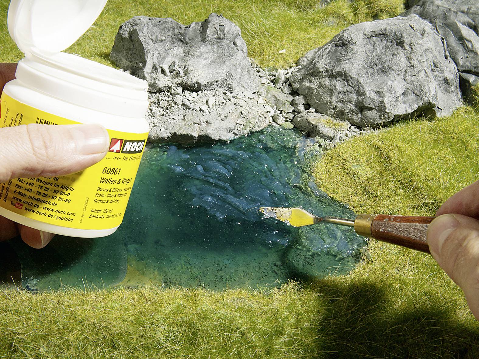 Osoba nakłada niebieską substancję narzędziem na sztuczny krajobraz z skałami i trawą.