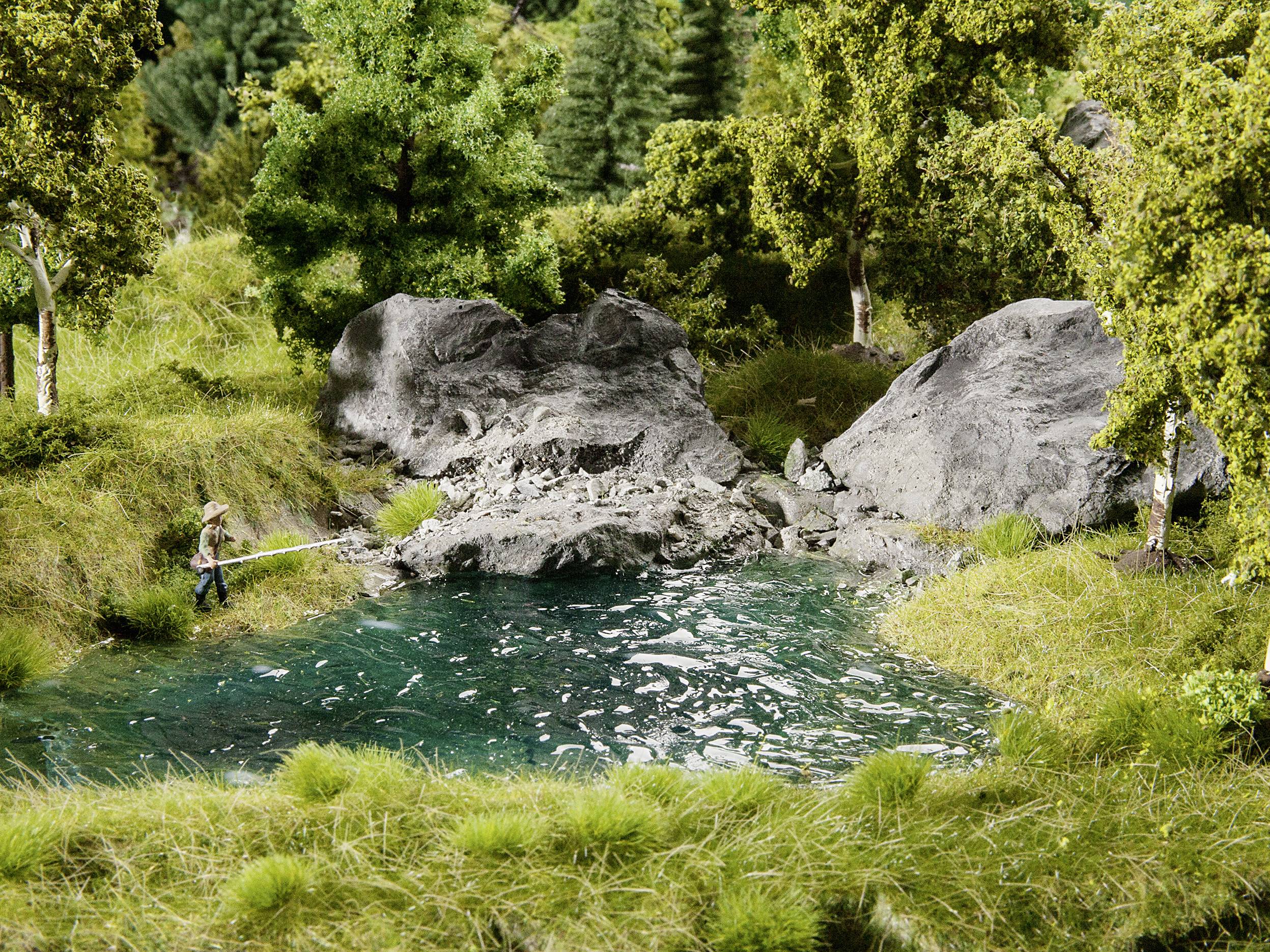 Mały staw w zalesionym krajobrazie skalistym. Jedna osoba wędkuje nad brzegiem, otoczona zielonymi drzewami i trawą.