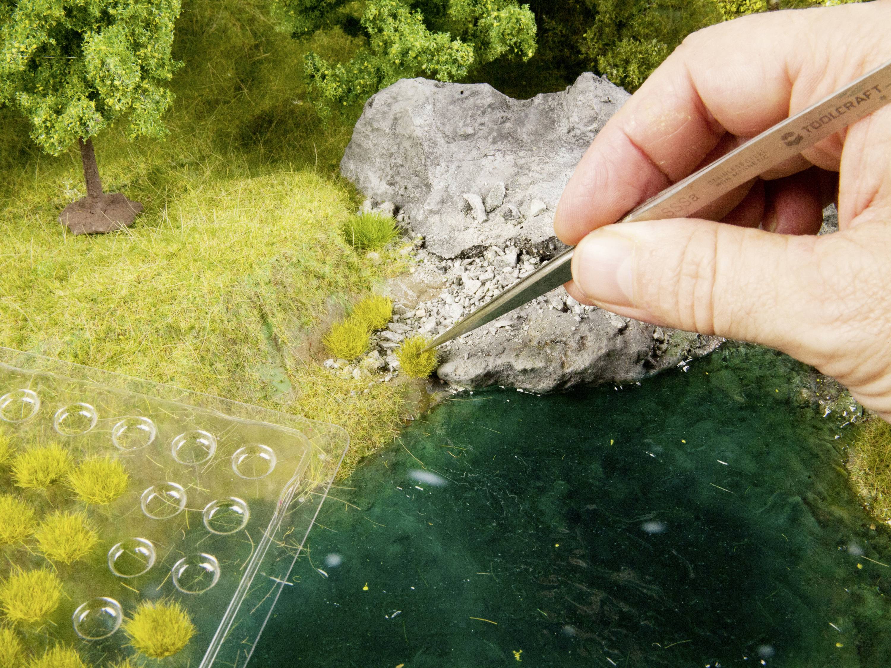 Dłoń umieszcza za pomocą pęsety małe kępy trawy nad modelowanym potokiem, w tle widoczne są skały i drzewa.