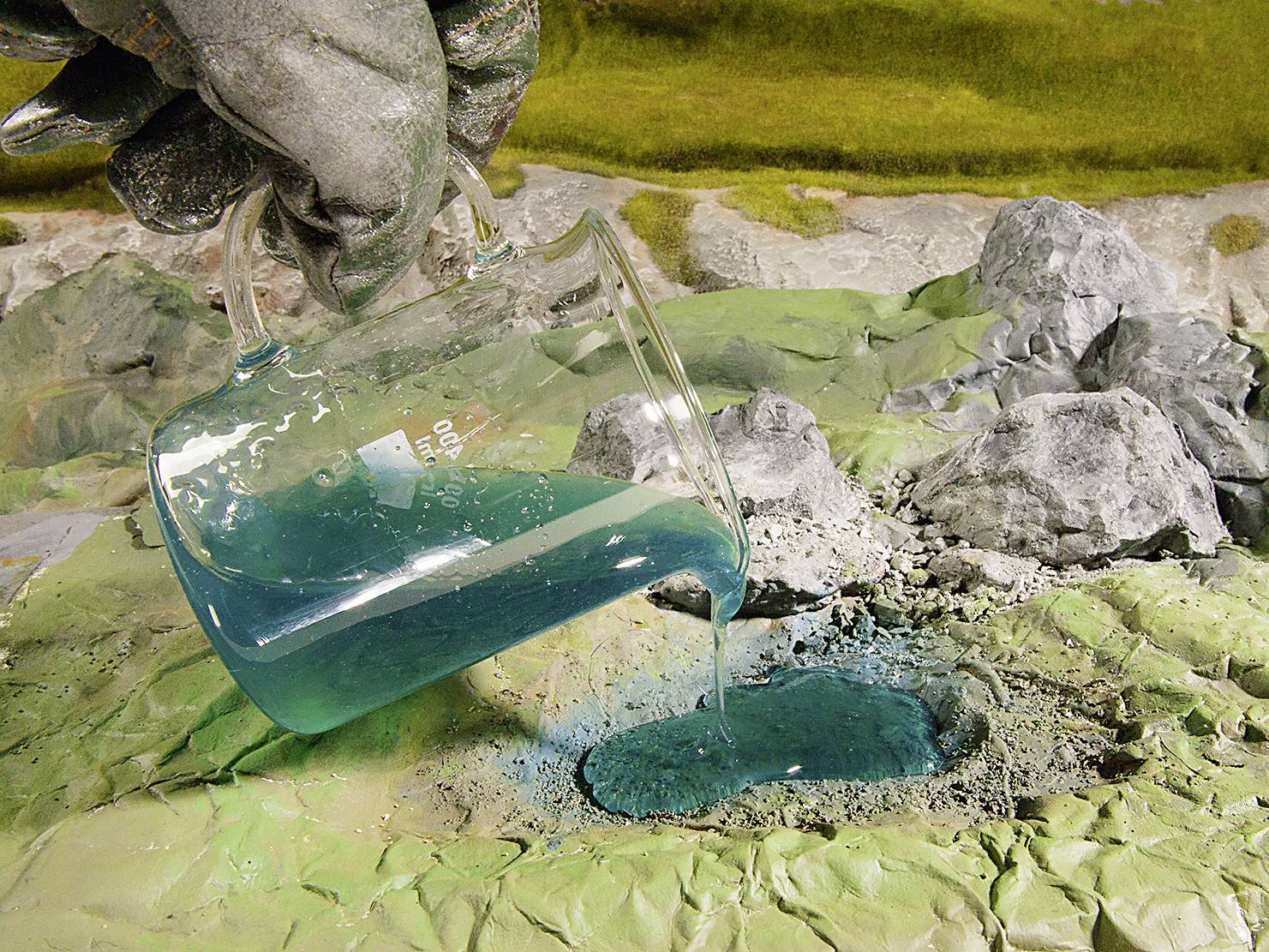 Dłoń w rękawicy ochronnej wylewa niebiesko-zieloną ciecz z zlewki na skalistą, zieloną powierzchnię. Eksperyment chemiczny.