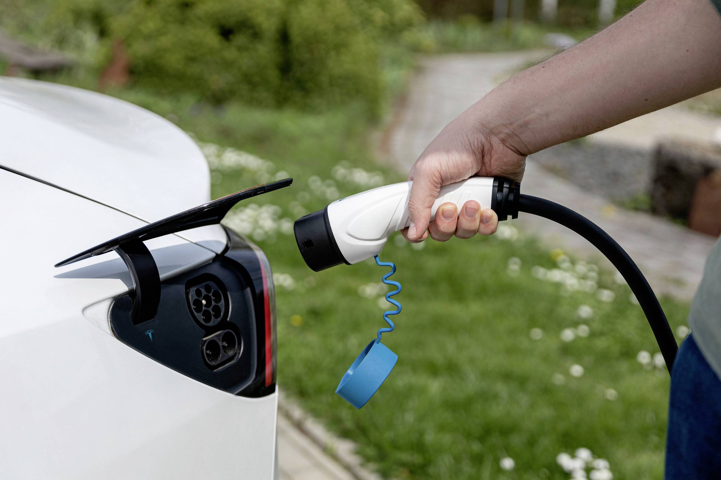 Osoba trzyma kabel ładujący i ładuje białego elektrycznego samochodu na wolnym powietrzu. W tle widoczne są tereny zielone.
