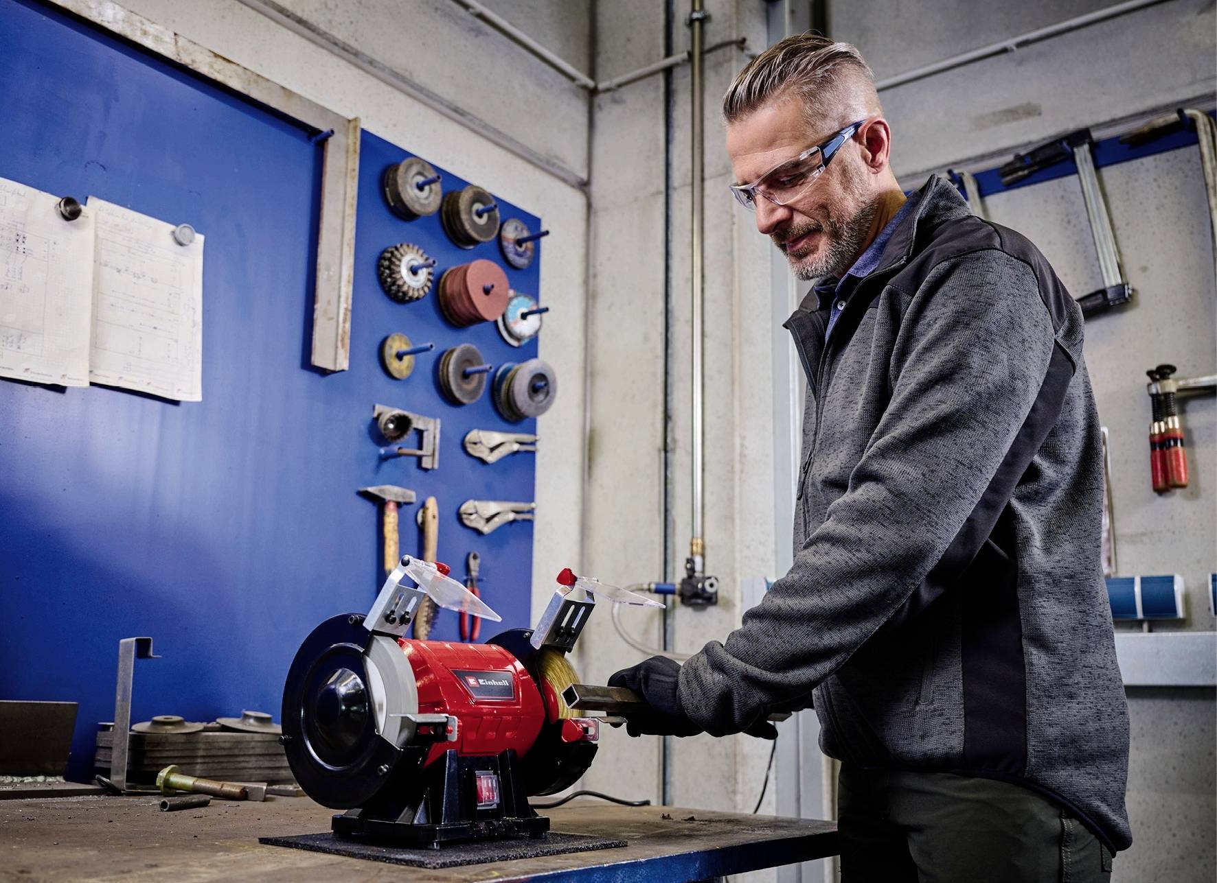 Mężczyzna obrabia narzędzie przy pomocy urządzenia szlifierskiego w warsztacie. Na ścianie wiszą narzędzia i tarcze szlifierskie.