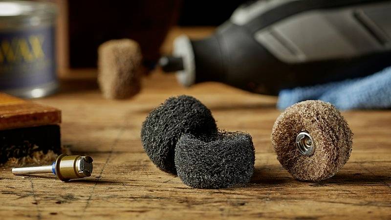 Różne nasadki szlifierskie leżą na drewnianym stole przed elektronarzędziem. W tle widoczna jest niebieska struktura tkaniny.