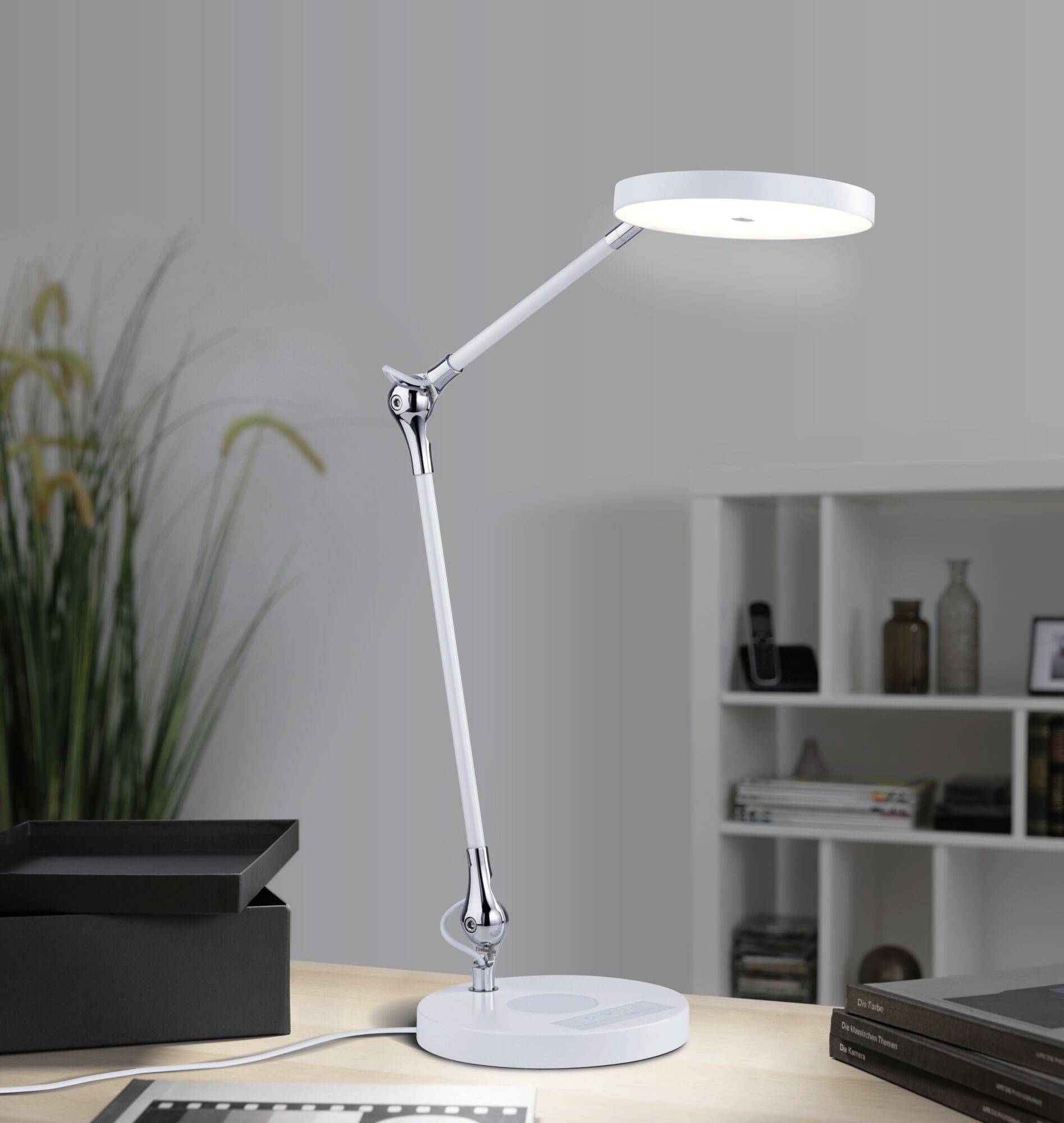 Nowoczesna lampa biurkowa na stole, świecąca jasnym światłem. W tle regał z książkami i roślina.