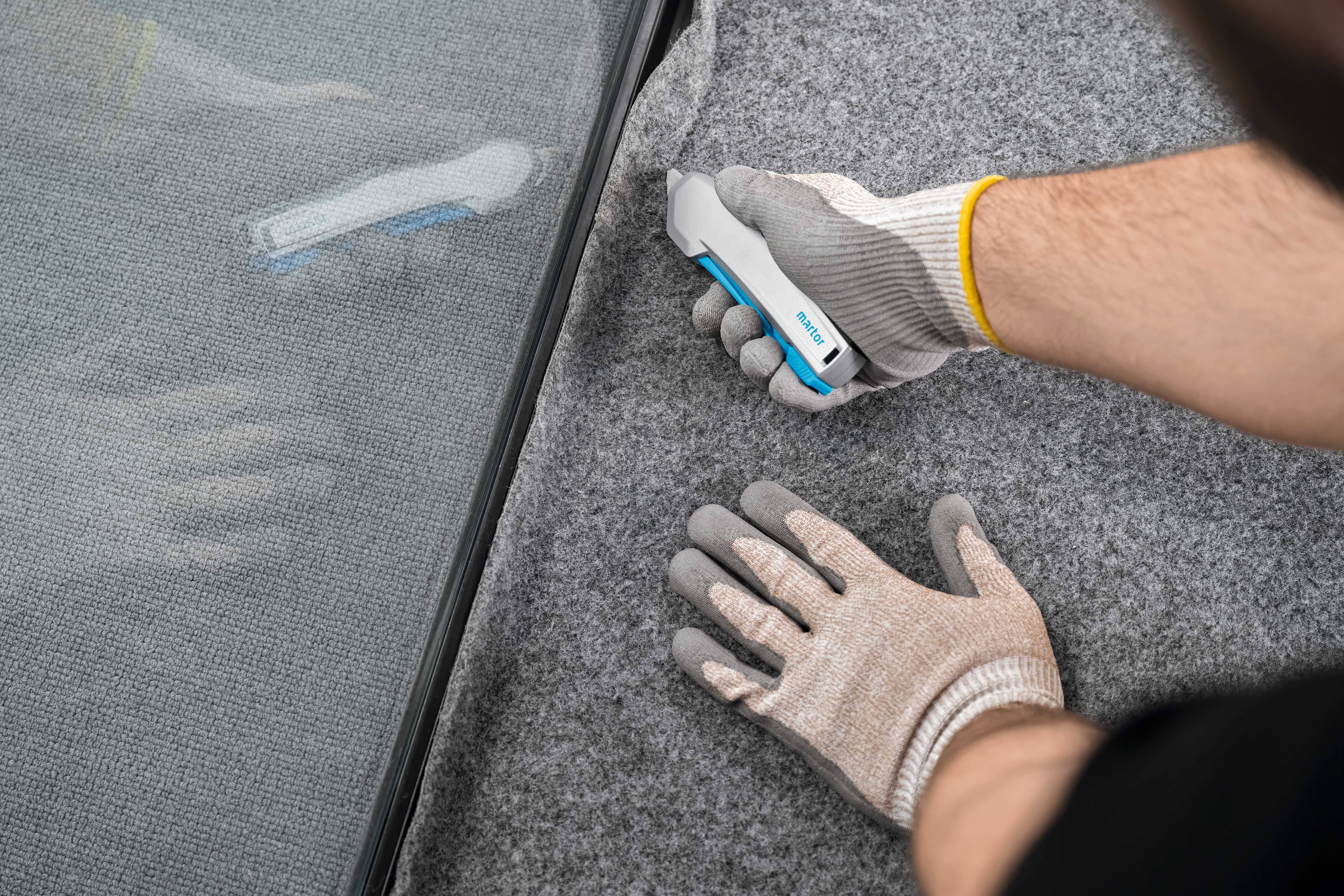 Osoba podcina szary dywan nożem do tapet w pobliżu krawędzi szkła. Ręce są zabezpieczone rękawicami roboczymi.