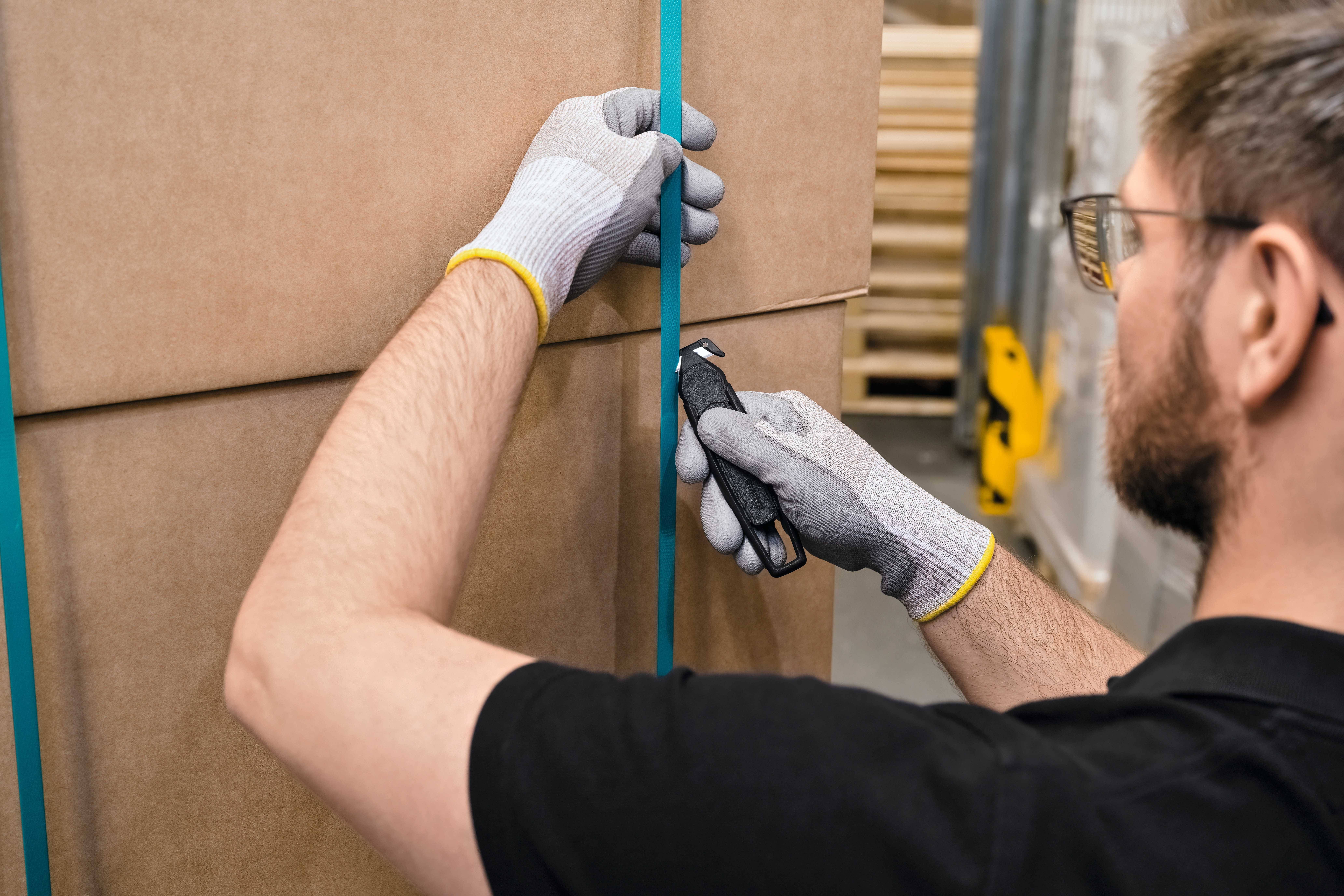 Mężczyzna w rękawiczkach przecina taśmę pakową na dużym kartonie. W tle widoczne są regały.