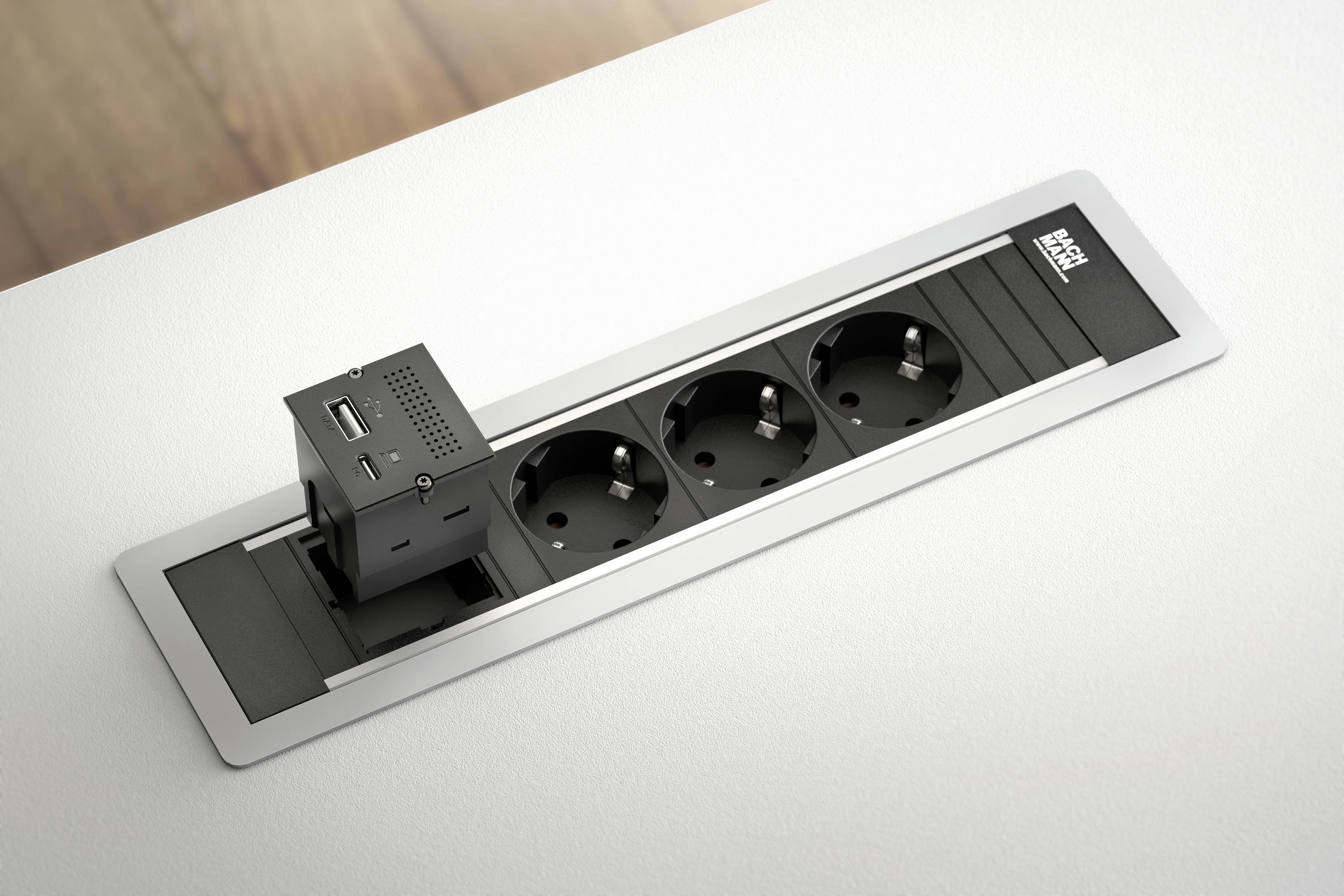 Wmontowany w blat stołowy moduł gniazdowy z trzema gniazdami elektrycznymi oraz adapterem do podłączenia USB, otoczony elegankim srebrnym ramnikiem.