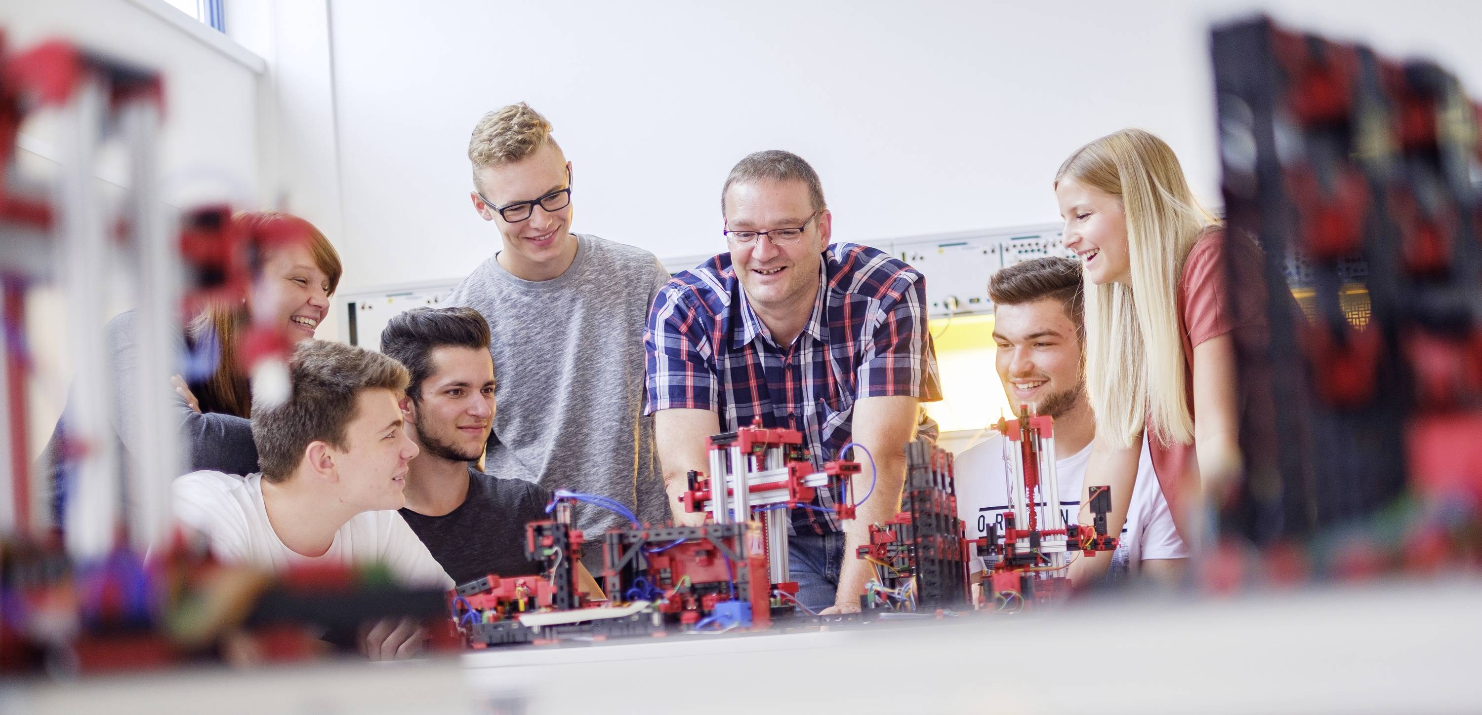 Grupa sześciu nastolatków śmieje się i wspólnie pracuje nad projektem technicznym z modelami robotów w jasnym pomieszczeniu.