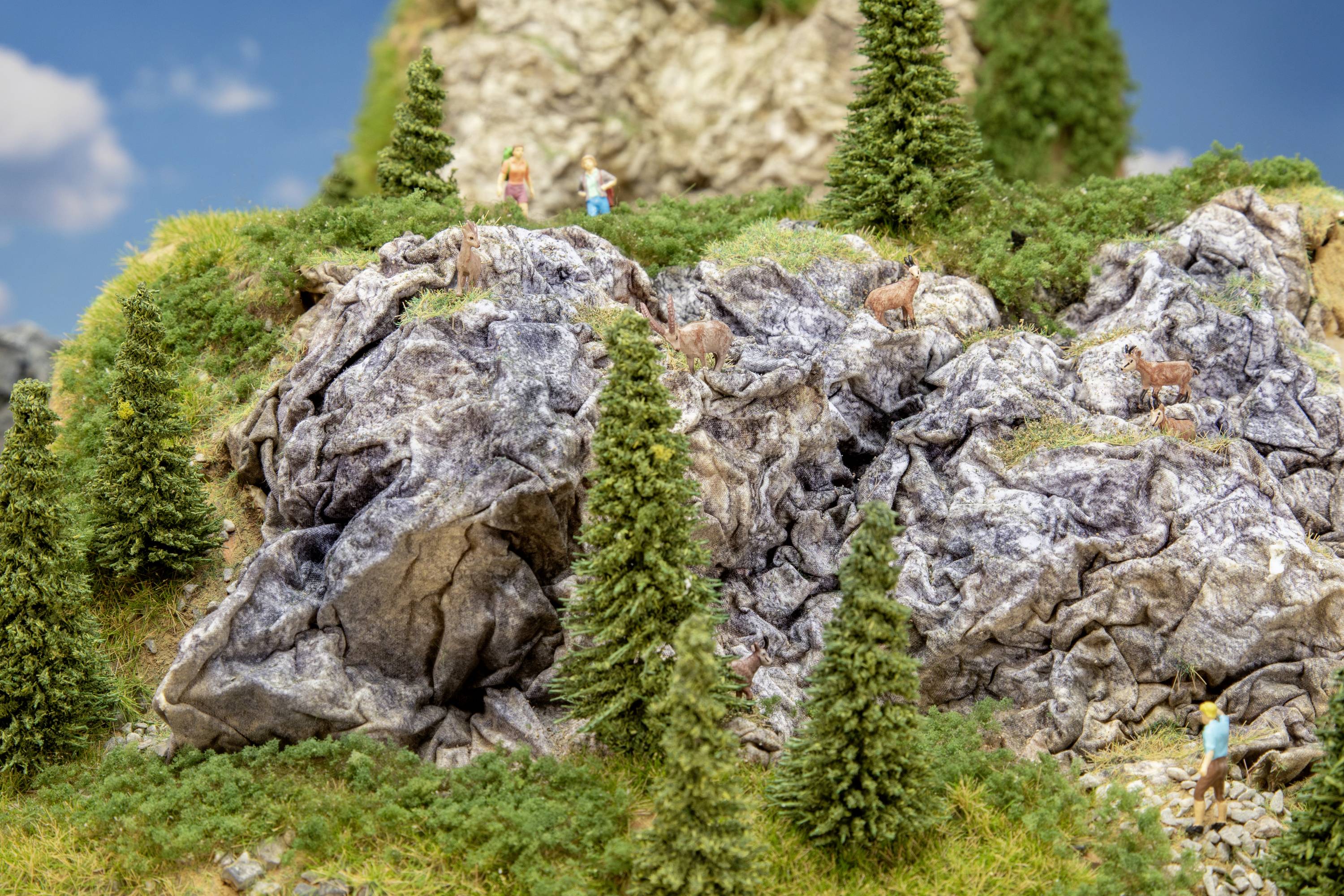 Miniaturowa sceneria przedstawia skalistą scenerię górską z małymi postaciami, świerkami i trawiastymi połaciami.