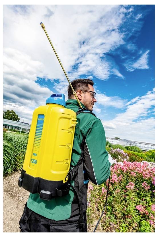 Osoba ubrana w zielony strój nosi żółty plecak natryskowy, idąc ścieżką ogrodową między kwitnącymi roślinami pod błękitnym niebem.