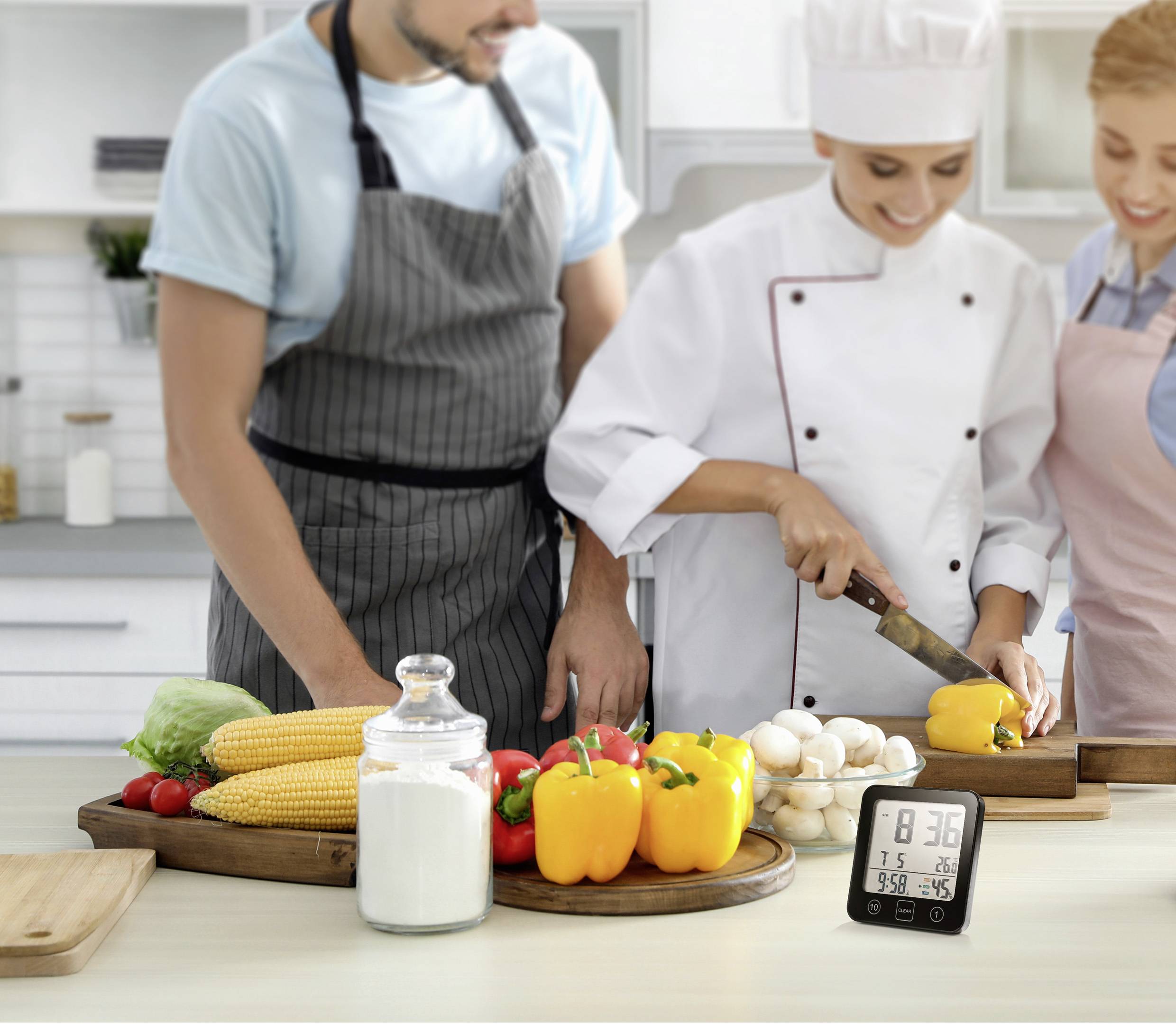 Kucharka kroi żółtą paprykę w nowoczesnej kuchni, stojąc obok dwóch osób. Na stole warzywa, przyprawy i minutnik kuchenny.
