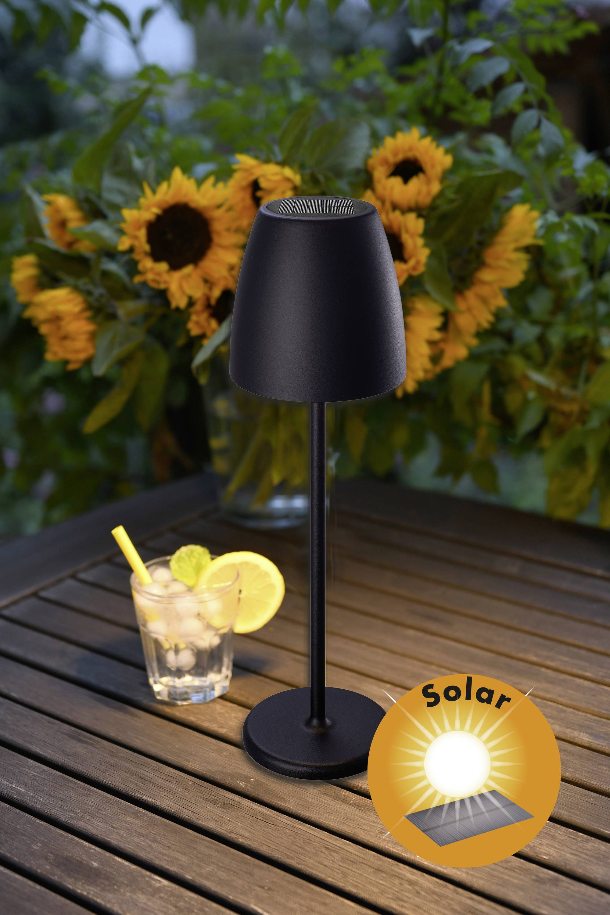 Czarna lampa solarna stoi na drewnianym stole przed bukietem słoneczników. Obok znajduje się szklanka z wodą z lodem i cytryną.