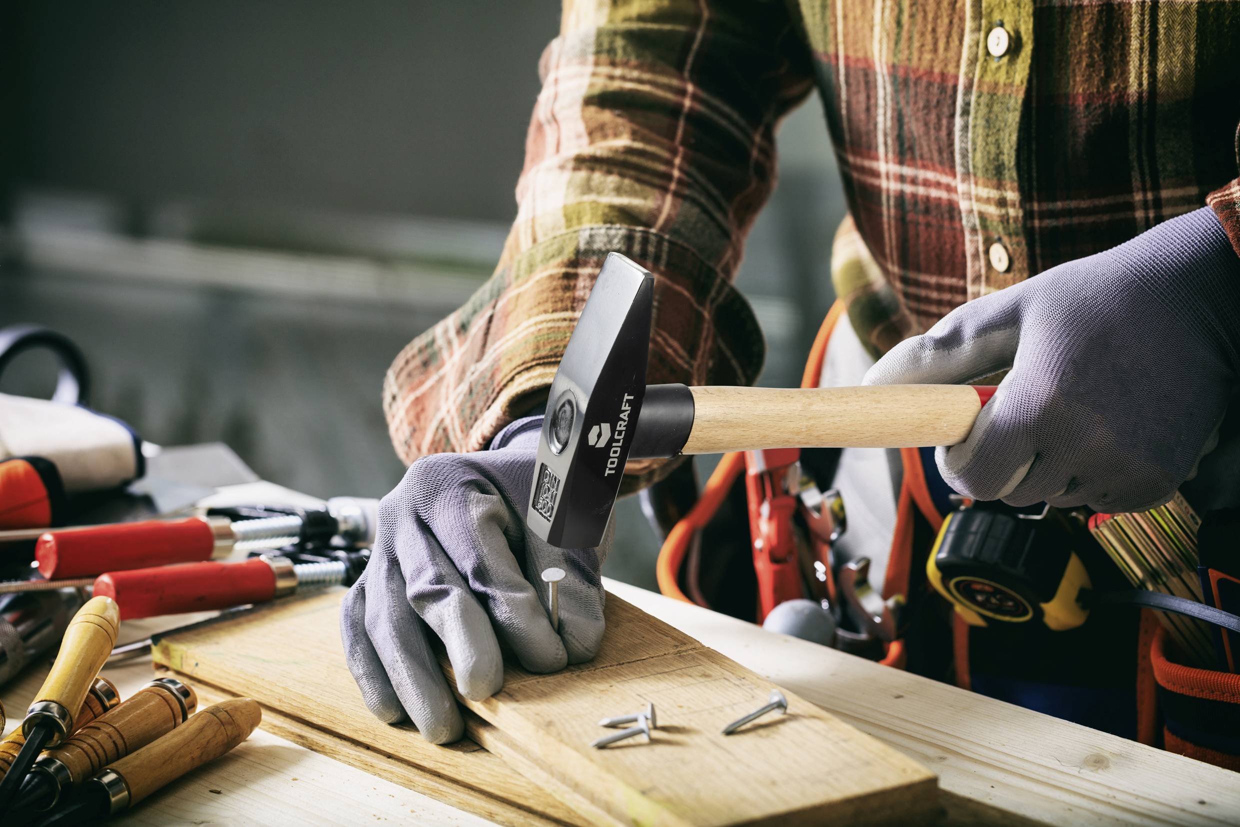 Osoba w kratowanej koszuli i rękawiczkach, która wbija gwoździa młotkiem w deskę drewnianą, otoczona narzędziami.