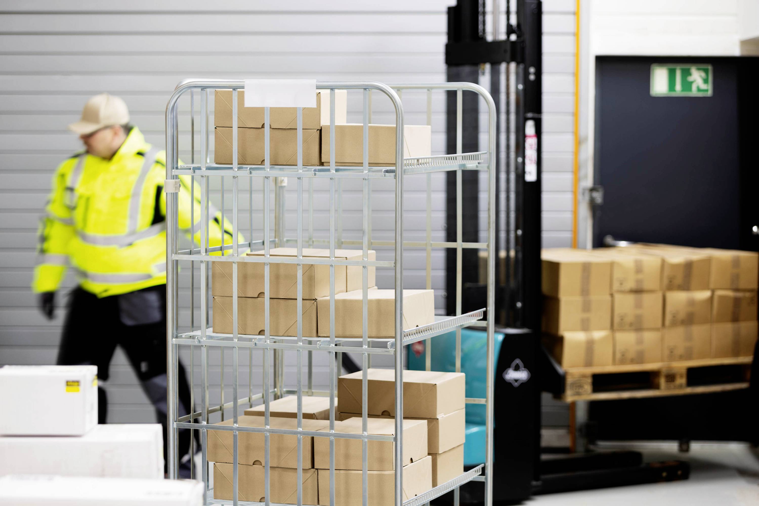 Robotnik w żółtej kurtce porusza się w magazynie, gdzie skrzynie są ułożone na wózku oraz na półce. W tle widoczne są drzwi wyjściowe.