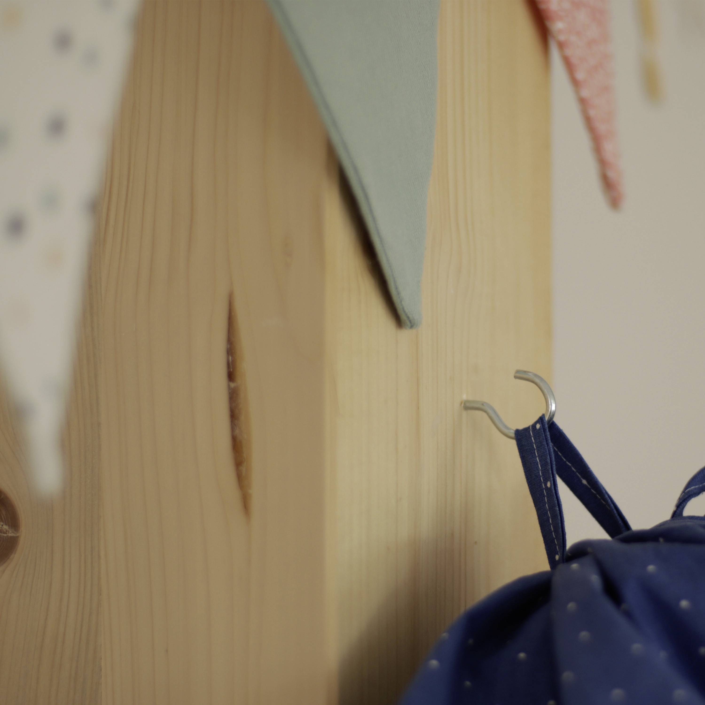 Niebieski kawałek tkaniny wisi na haczyku na drewnianej ścianie; ponad nim kolorowe trójkątne kawałki materiału utworzone są w girlandę.