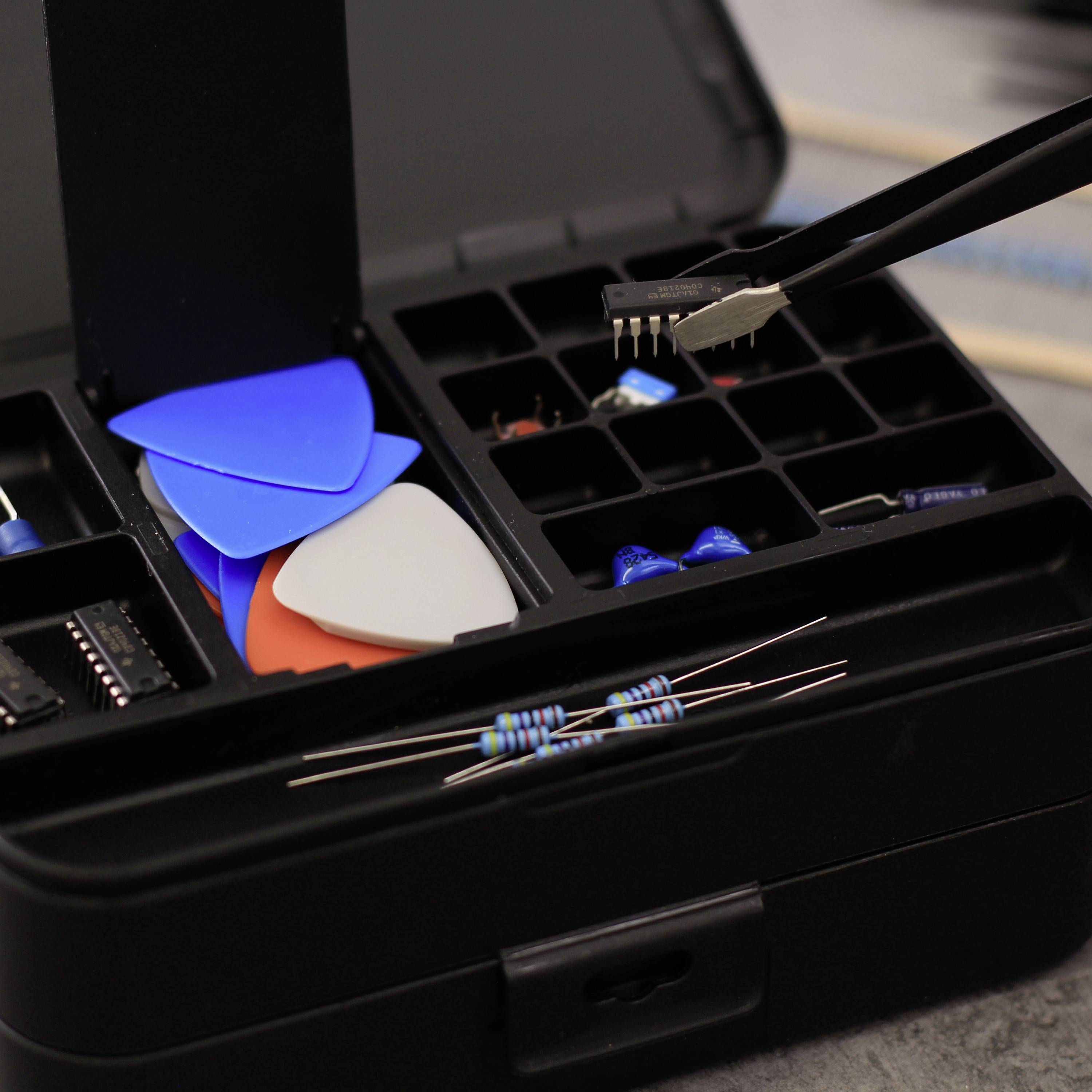 Otwarty przybornik z kostkami gitarowymi, elementami elektronicznymi takimi jak rezystory i mikroprocesor. Narzędzie trzyma mikroprocesor nad skrzynką.