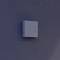 Biały, kwadratowy przycisk zamontowany jest na gładkiej, szarej ścianie. Urządzenie posiada dwa przyciski i małe logo w prawym górnym rogu.