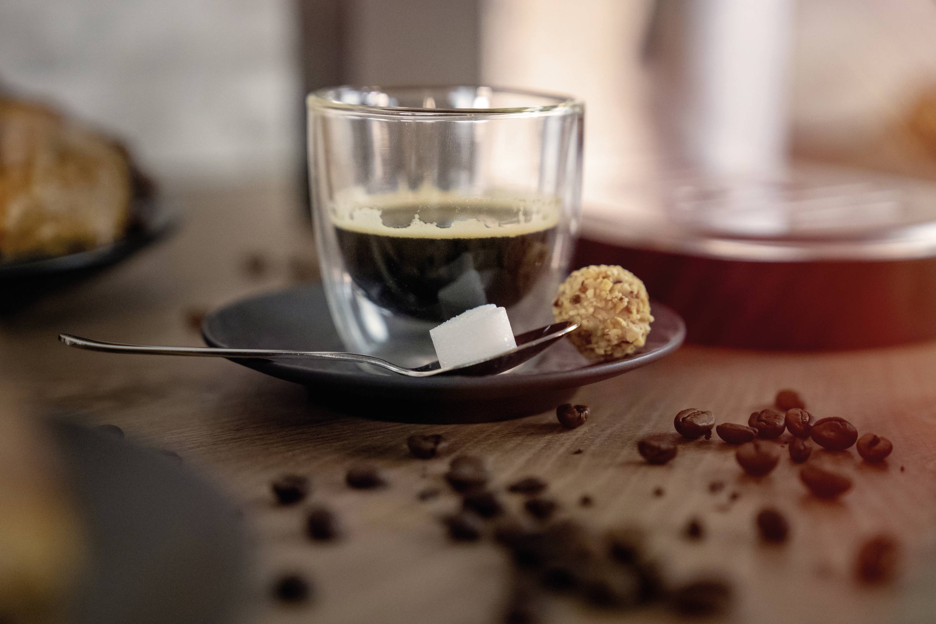 Filiżanka espresso na spodeczku, obok kostki cukru i wypieku. Na stole rozsypane ziarna kawy.