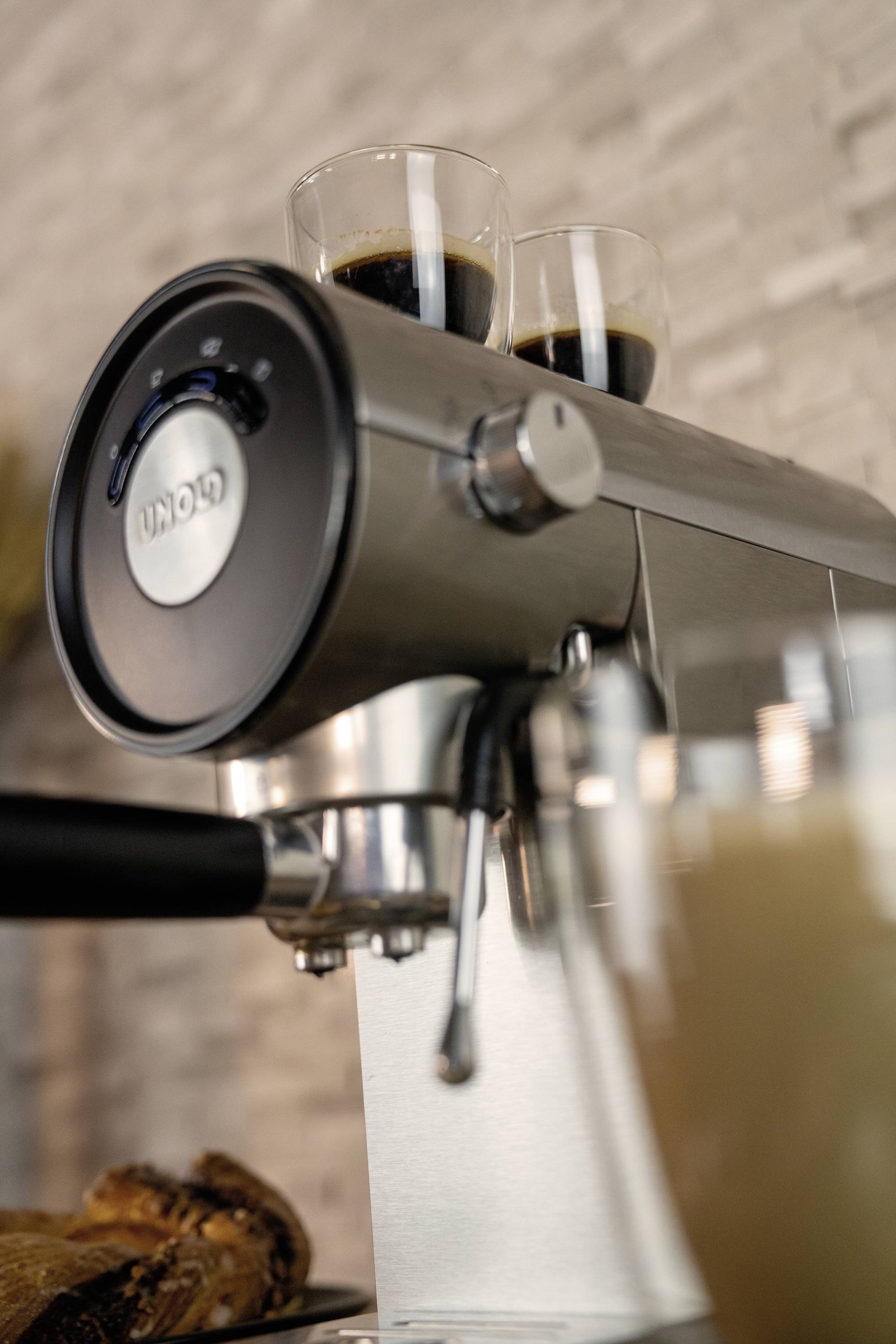 Ekspres do espresso z dwoma filiżankami espresso na wierzchu, w tle rozmyte wnętrze kawiarni.